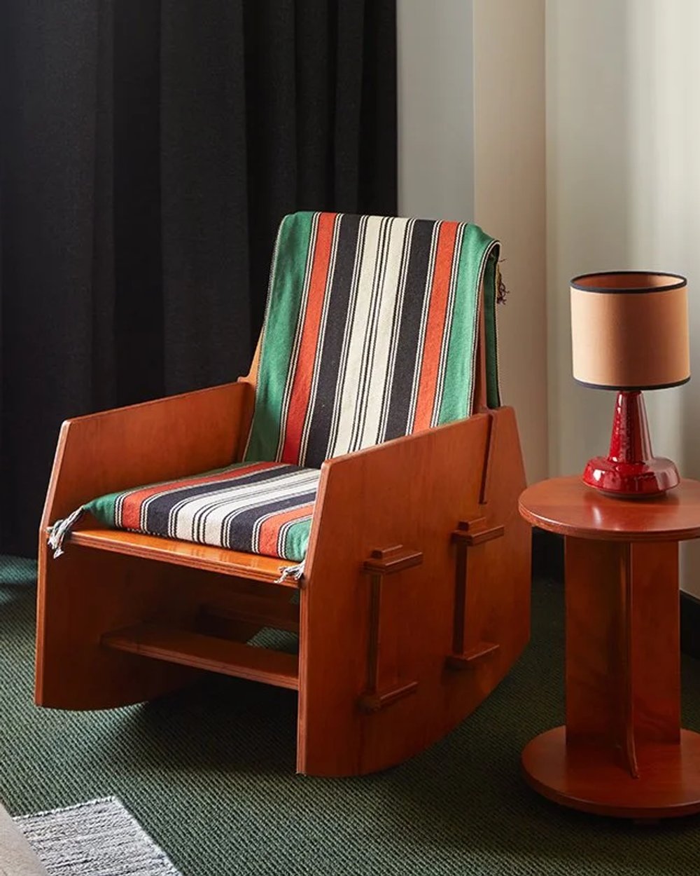 Hotel room corner with a cherry-red wooden rocking chair under a striped throw, beside a small red side table and lamp on green carpet