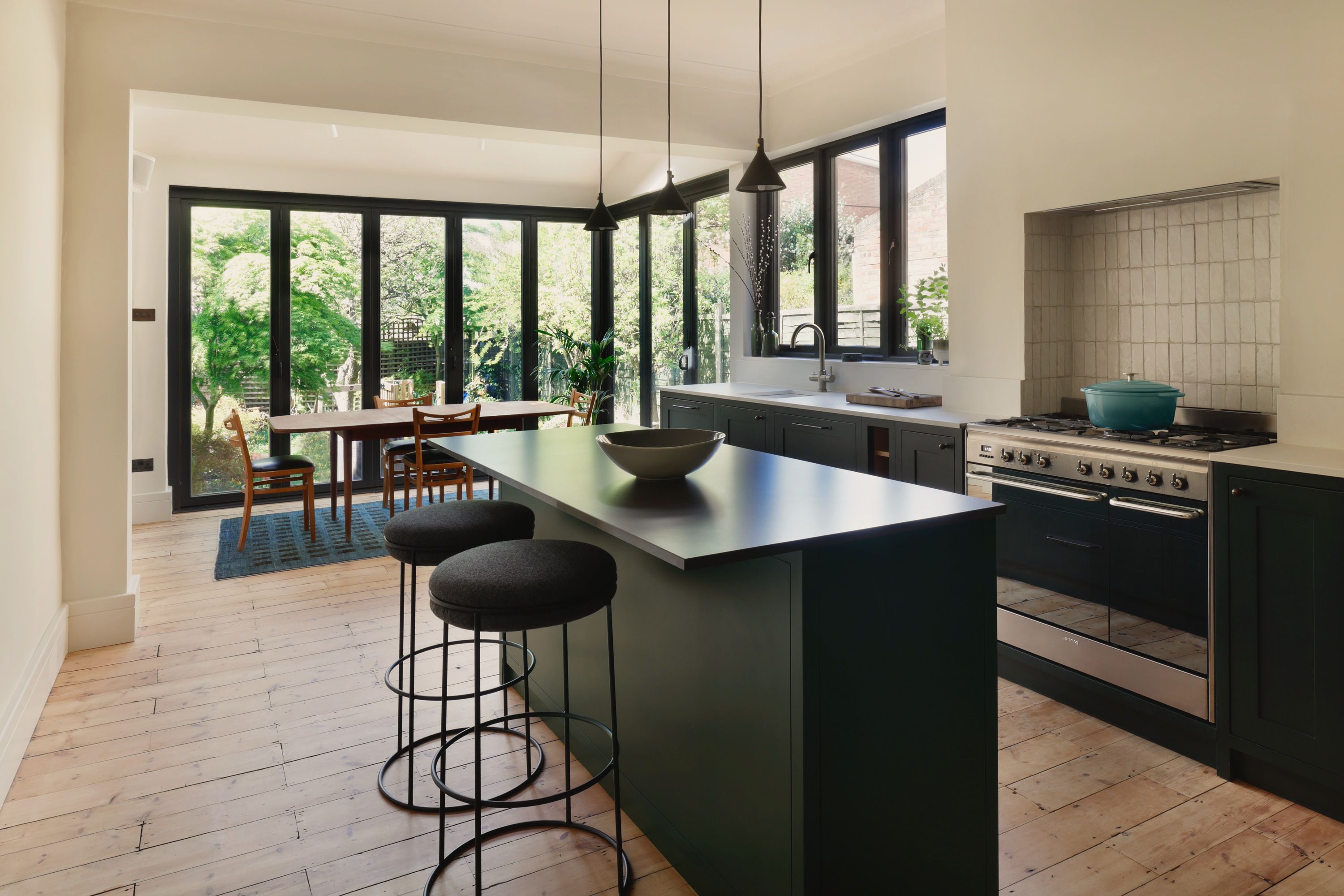 Dark green kitchen with a stone-topped island and two stools, a range cooker under a tiled recess, and the glazed rear extension opening onto the garden.