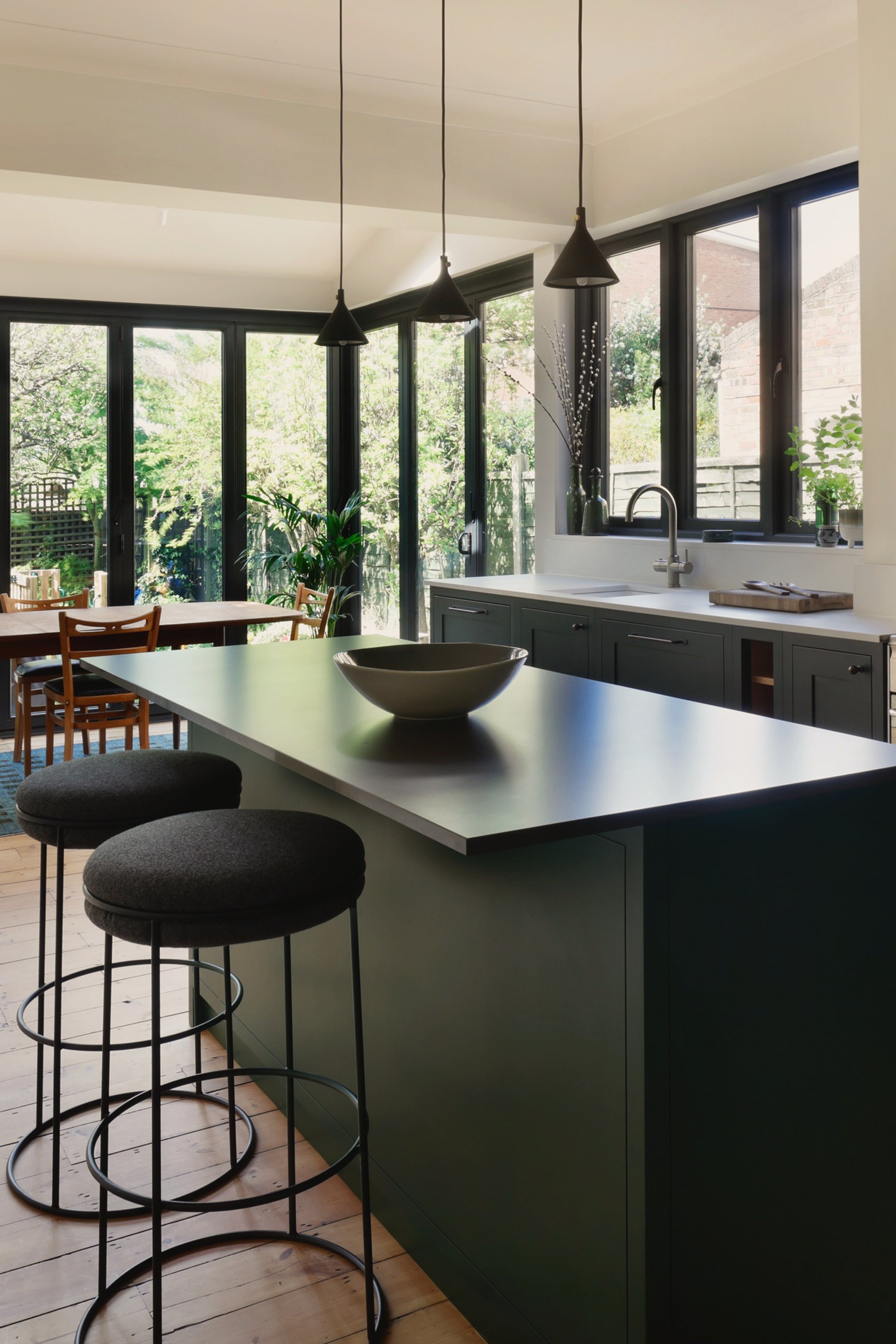 Dark green kitchen with a stone-topped island and two stools, a range cooker under a tiled recess, and the glazed rear extension opening onto the garden.