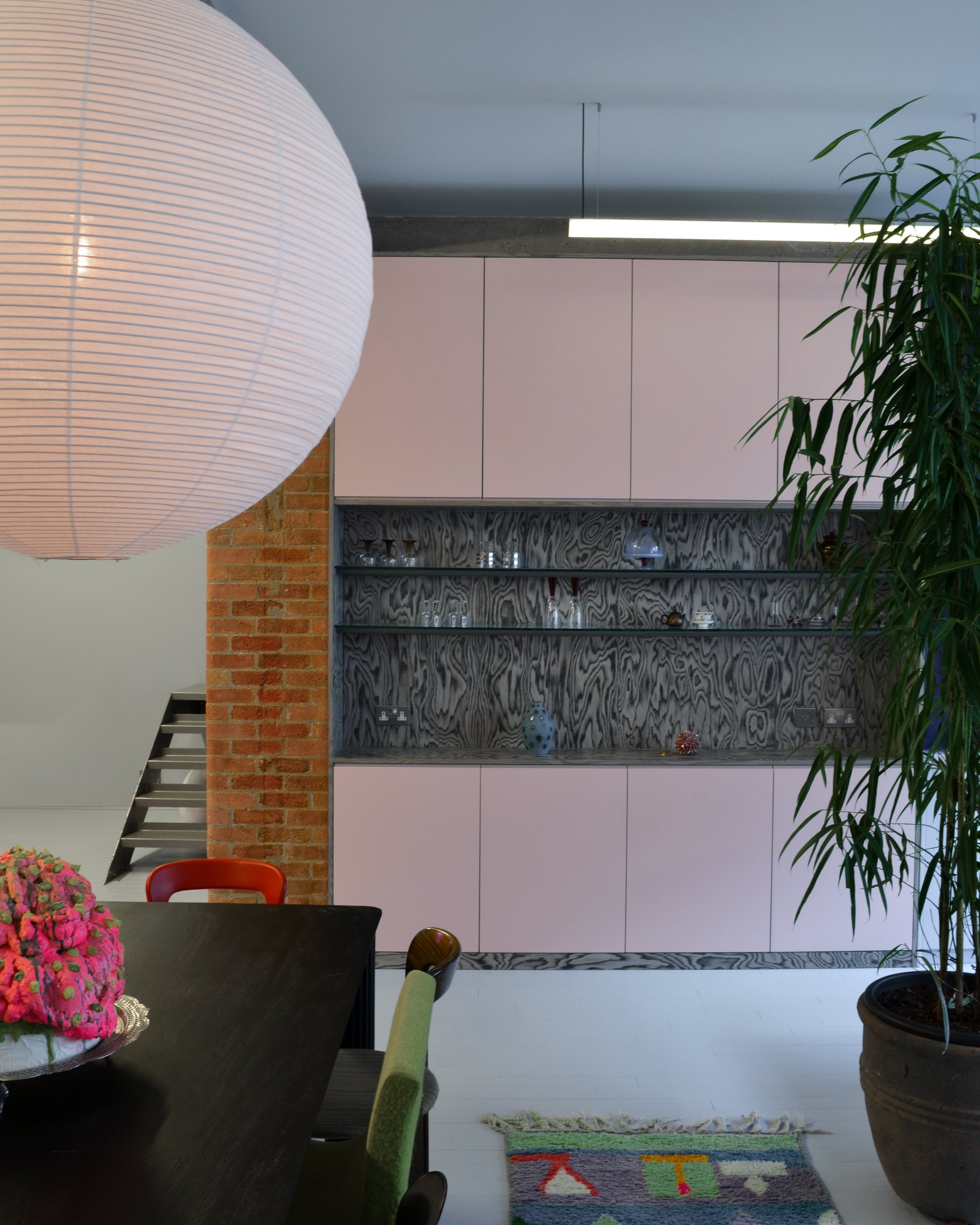 Dining area under a large pink paper globe, with pale pink cabinets, an antiqued mirror shelf of glassware and an exposed brick pier