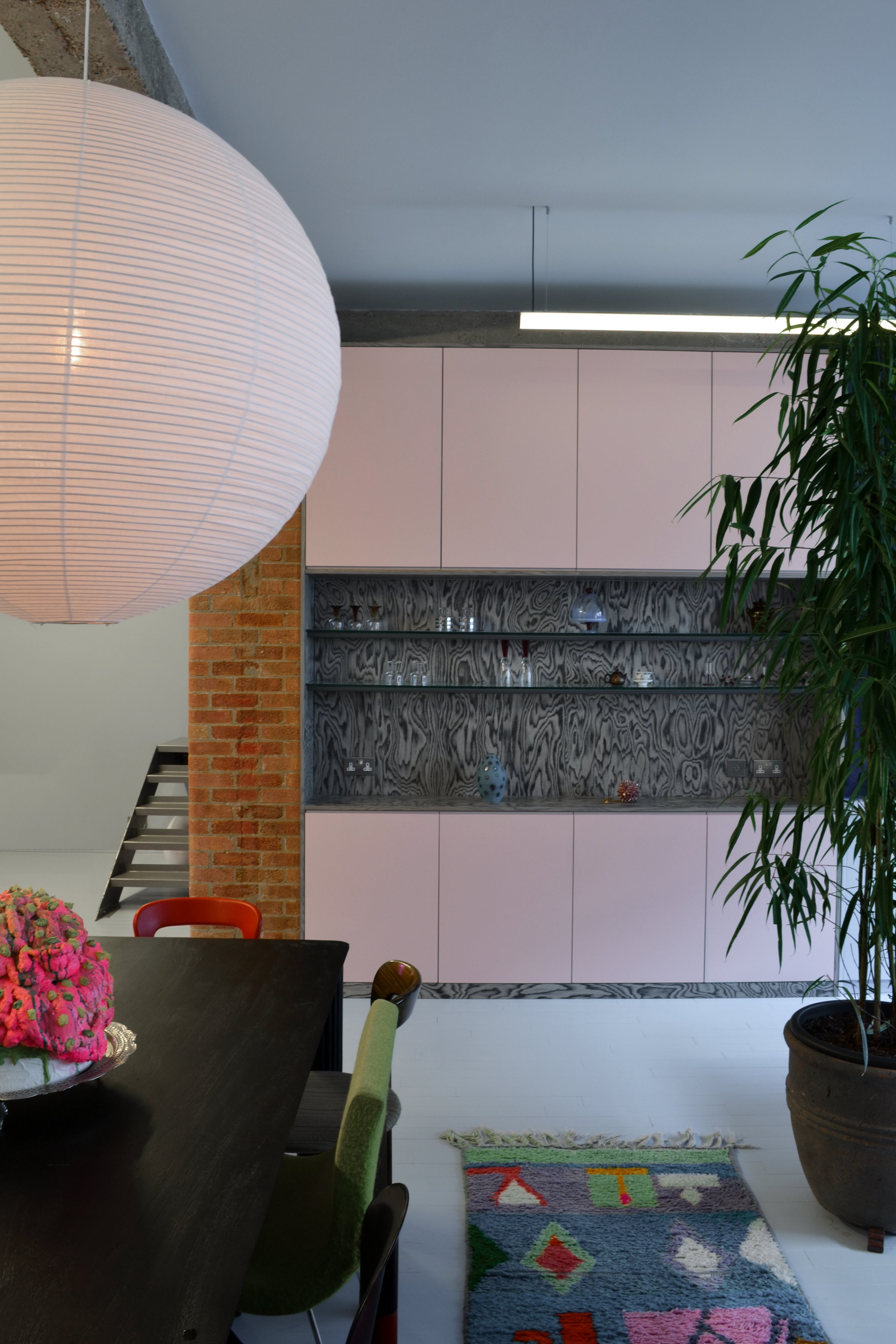 Dining area under a large pink paper globe, with pale pink cabinets, an antiqued mirror shelf of glassware and an exposed brick pier