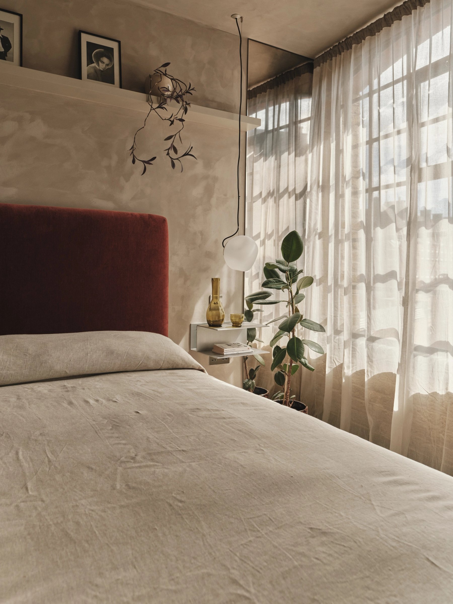 Bedroom from the foot of the bed, rust velvet headboard against polished plaster, a picture shelf above and sheer curtains filtering light.