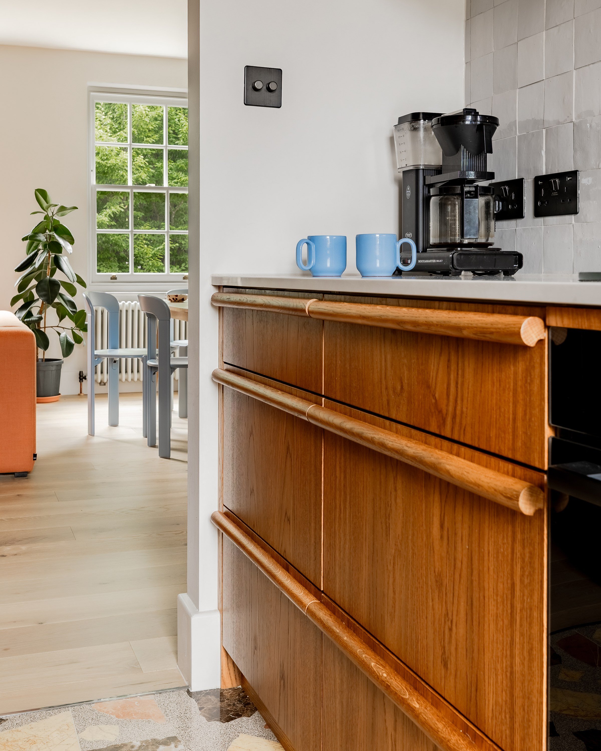 Wooden drawer fronts with long rounded pulls beside a coffee machine, the dining room bright beyond.