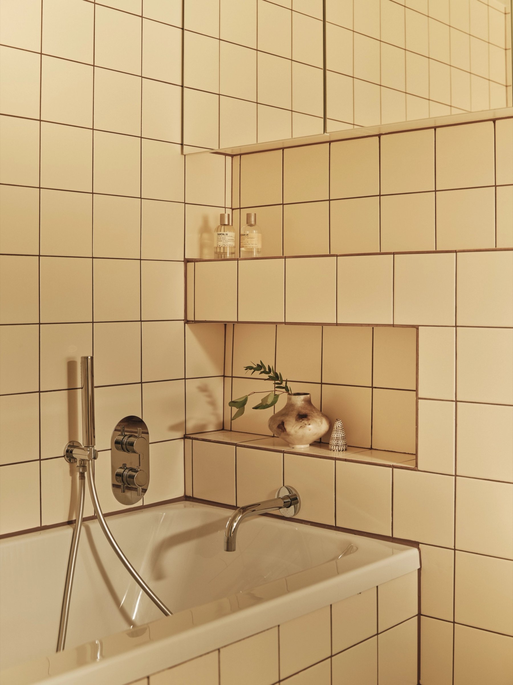Bathroom detail where square cream tiling steps out to form two shelves, one holding bottles and one a ceramic vase, above a chrome bath filler.