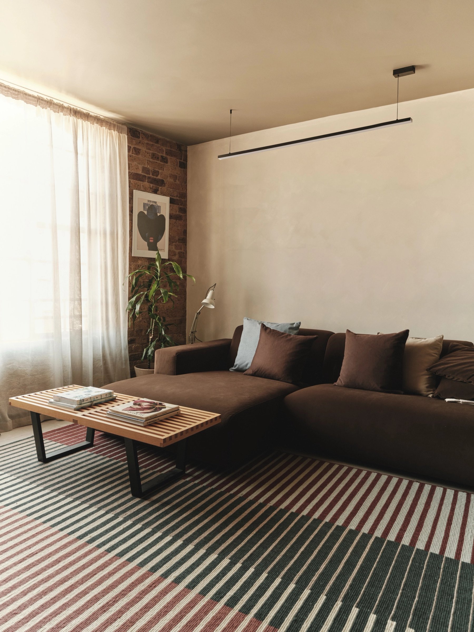 Sitting room with a deep brown corner sofa on a striped rug, a slatted timber coffee table and a framed poster against exposed brick.