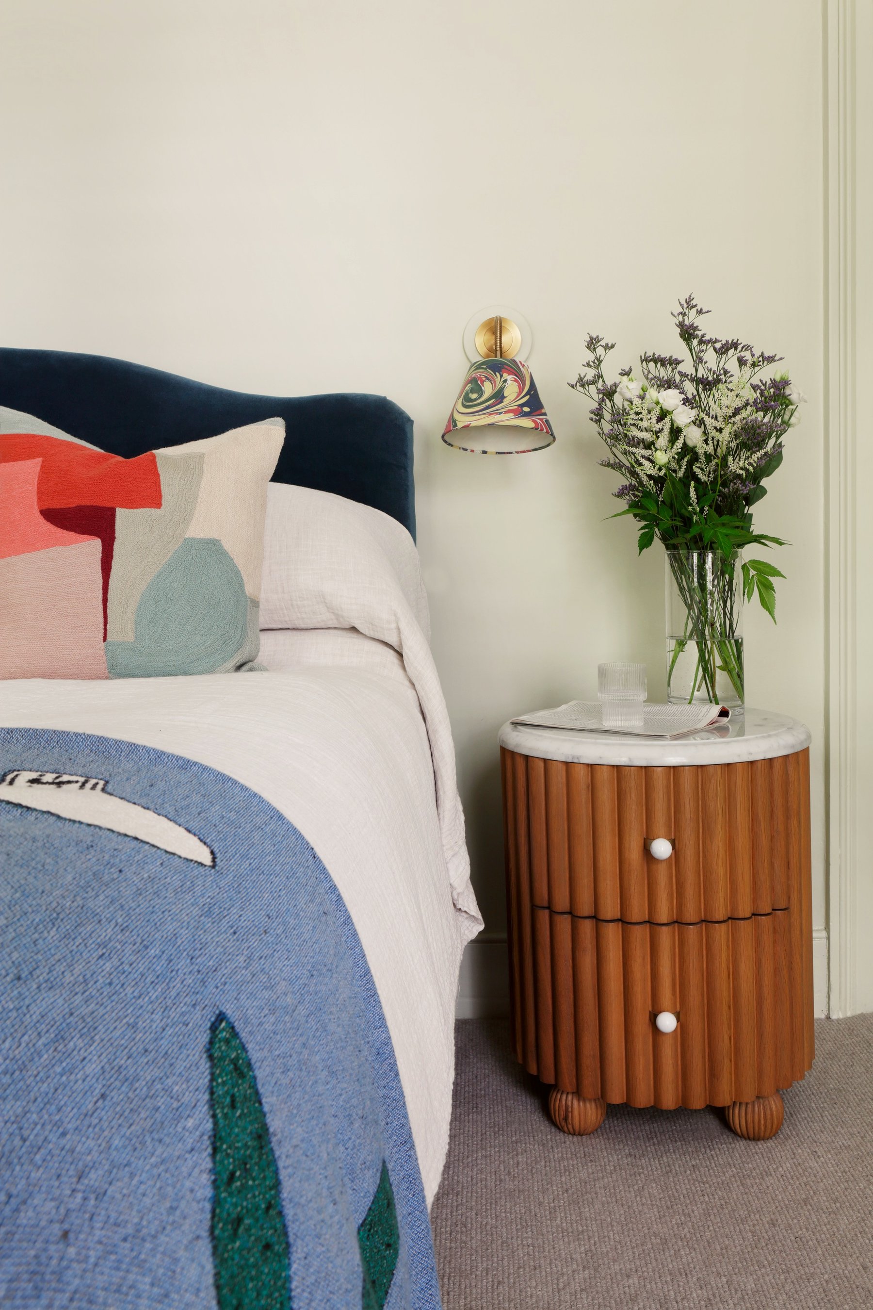 Bedside detail: a navy velvet headboard beside a fluted timber bedside table with a marble top, under a brass wall light with a painted glass shade.