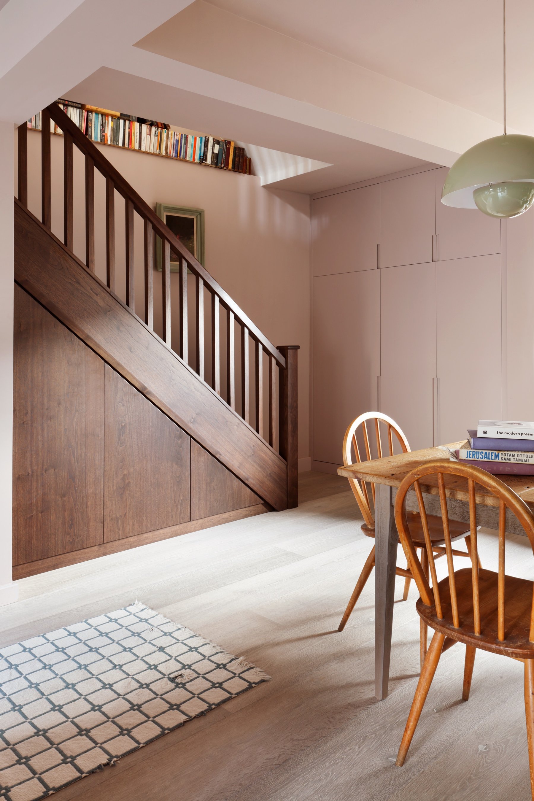 Dining end of the kitchen, dark timber stair and under-stair storage left, pale cupboards behind, a timber table under a green glass pendant.