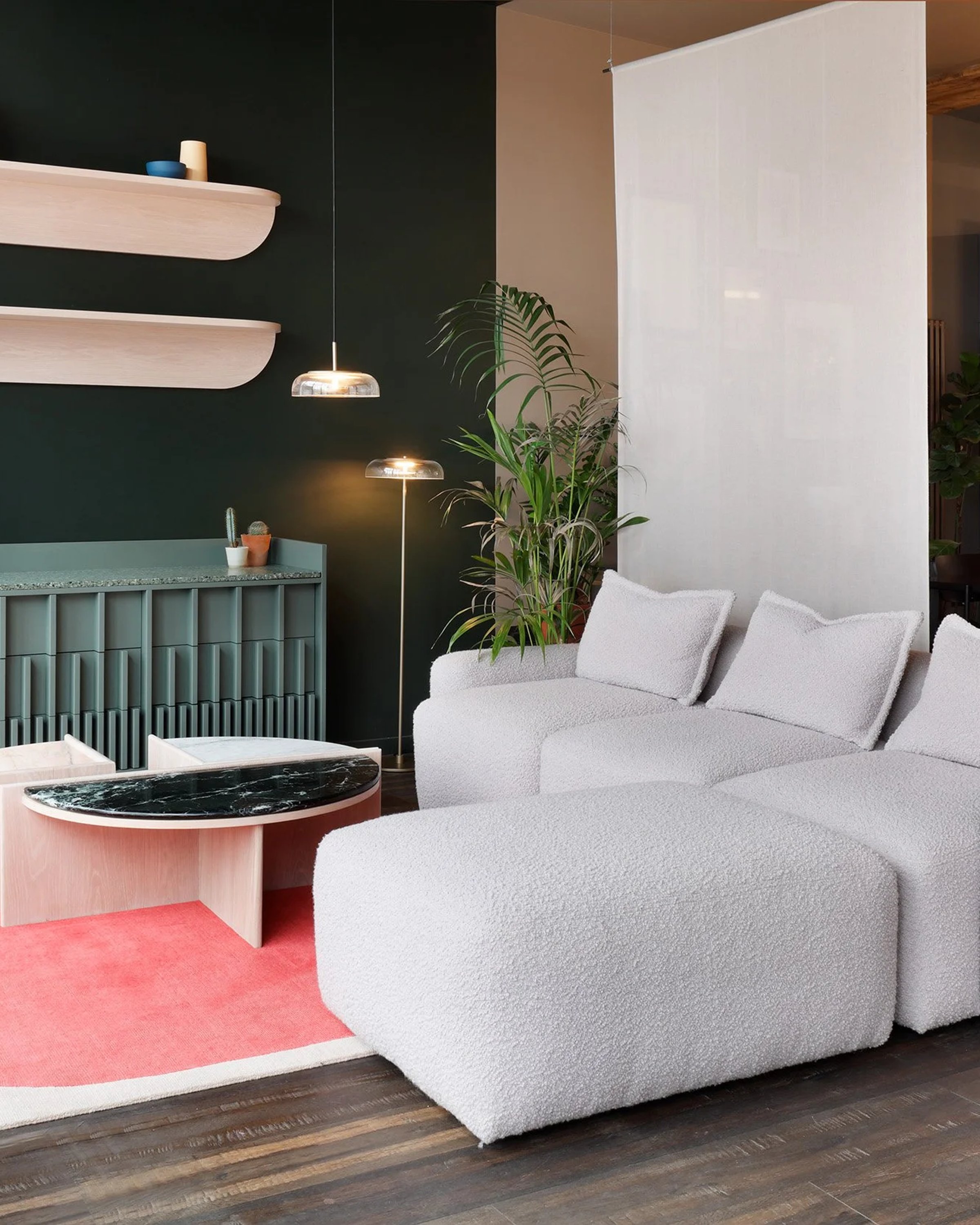 Showroom with a dark green wall, two curved pink floating shelves above a ribbed green sideboard, and a white boucle modular sofa on a round pink rug