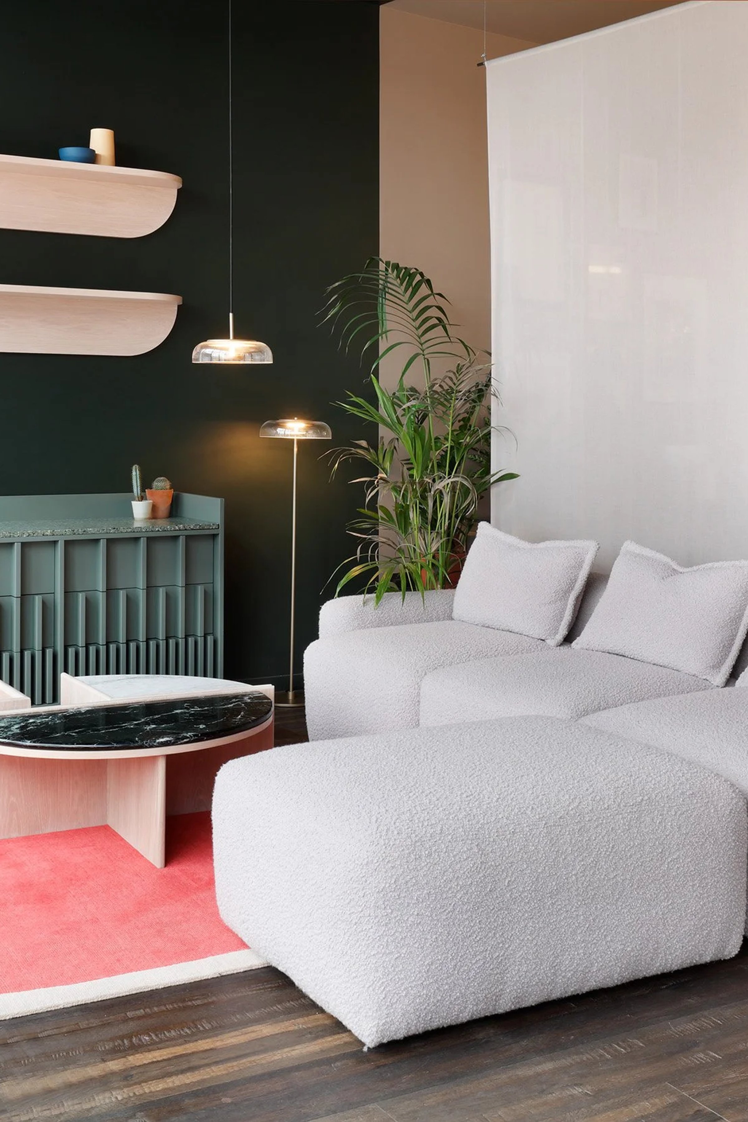 Showroom with a dark green wall, two curved pink floating shelves above a ribbed green sideboard, and a white boucle modular sofa on a round pink rug