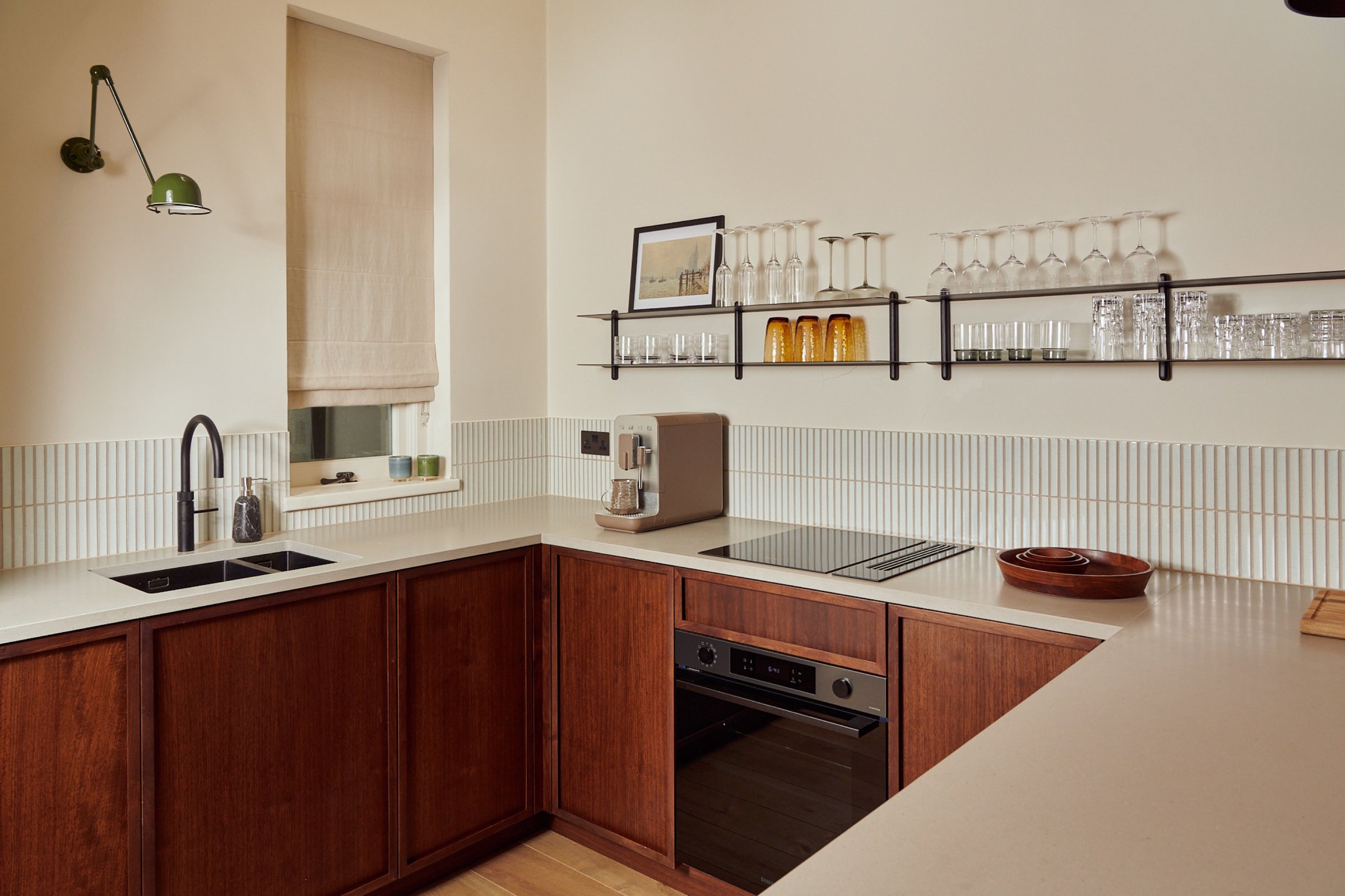 Kitchen corner in walnut with an integrated oven and hob, a fluted white splashback and glassware ranged on two black wall shelves.
