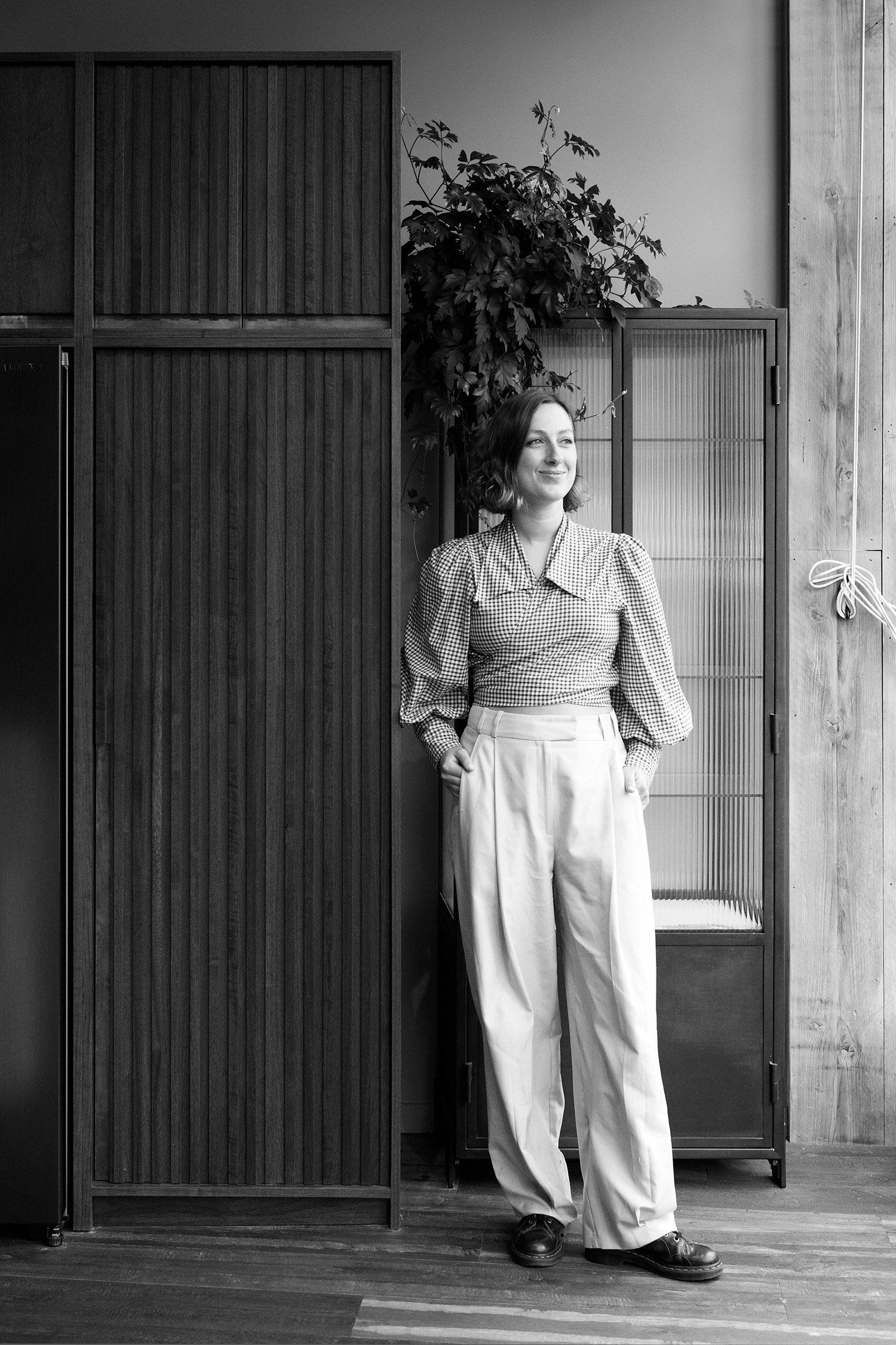 Angelica Smith in a gingham blouse and wide trousers, standing between dark slatted joinery and a glass cabinet.