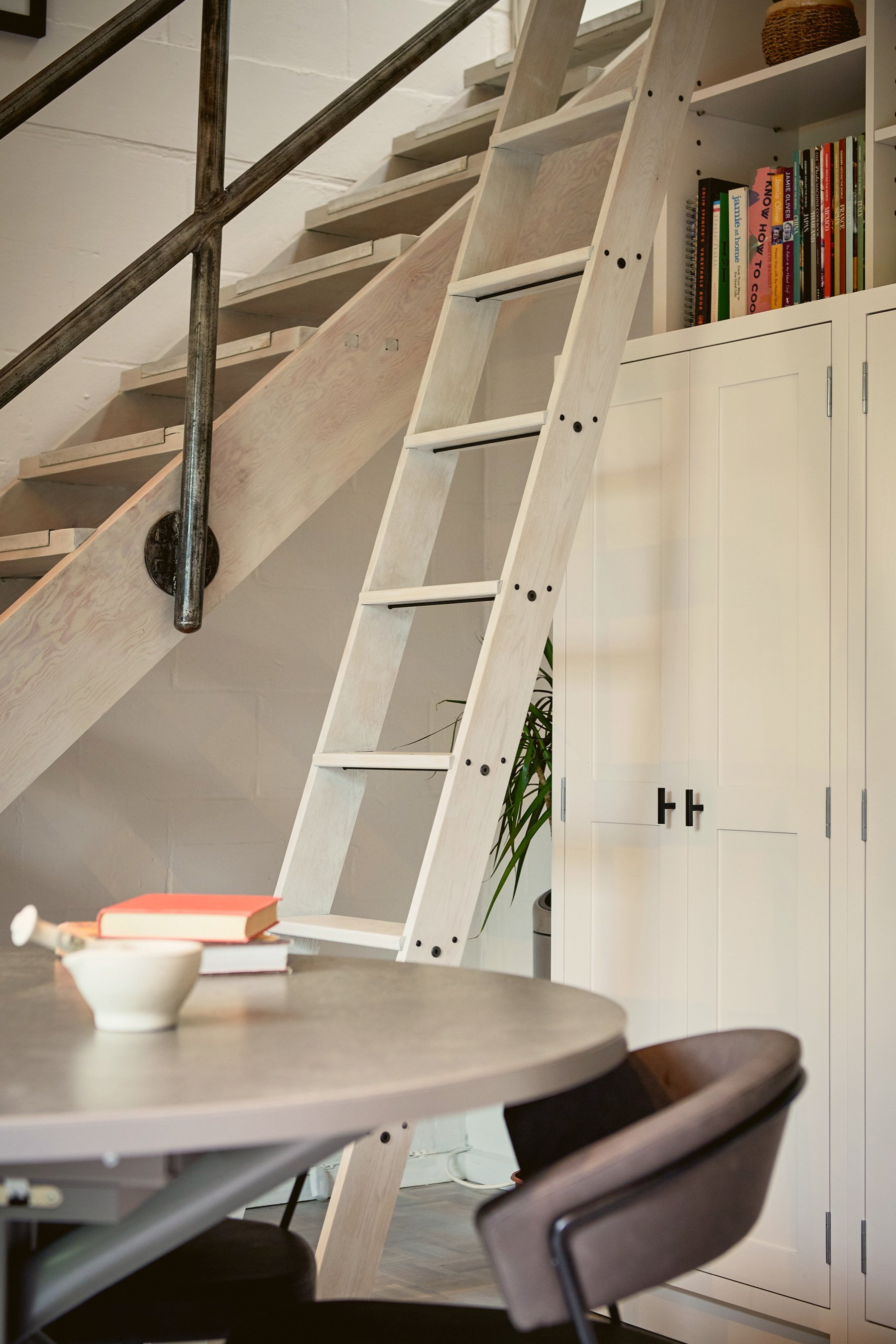 Corner beneath the stair, a limed timber library ladder leaning against the joinery beside an old iron handrail, with a round grey table in front.