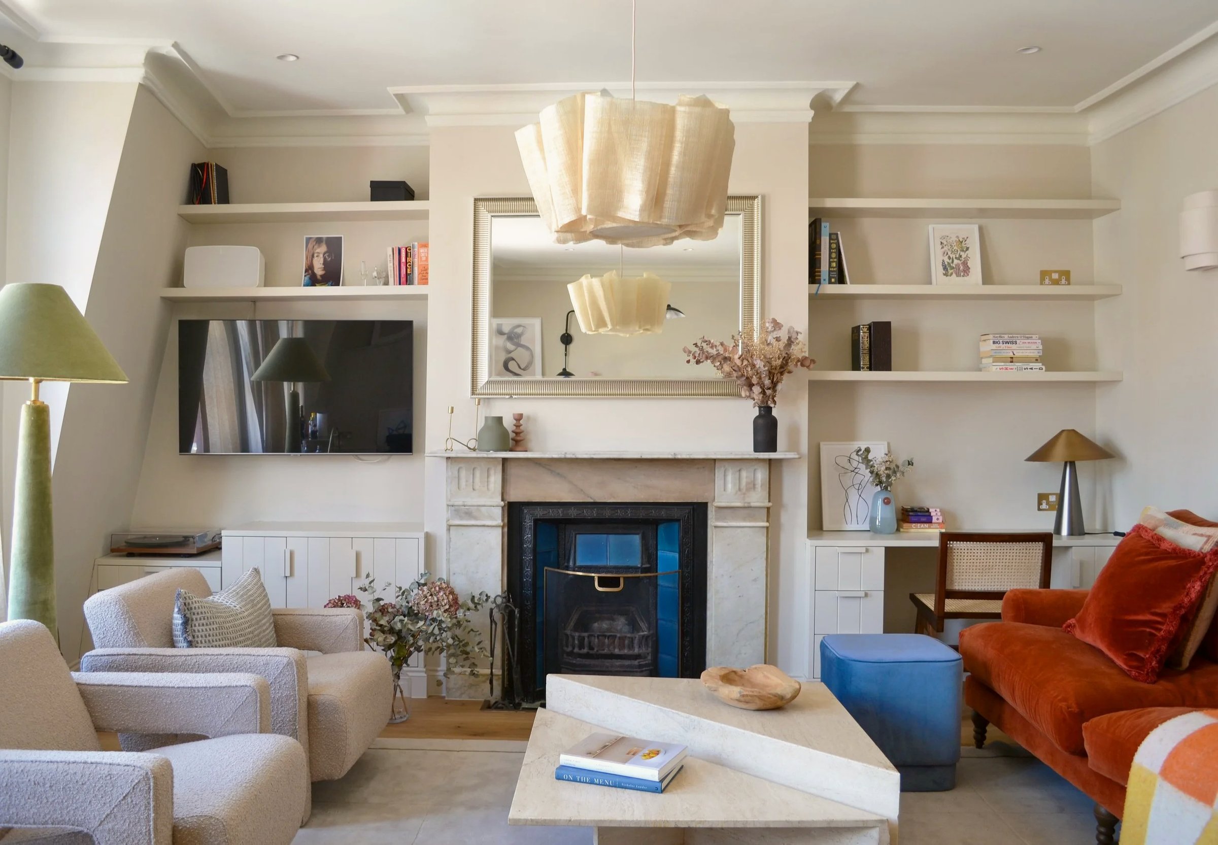 Living room with alcove shelving either side of a marble fireplace, a blue-tiled insert, pleated pendant light, burnt orange sofa and cream armchairs