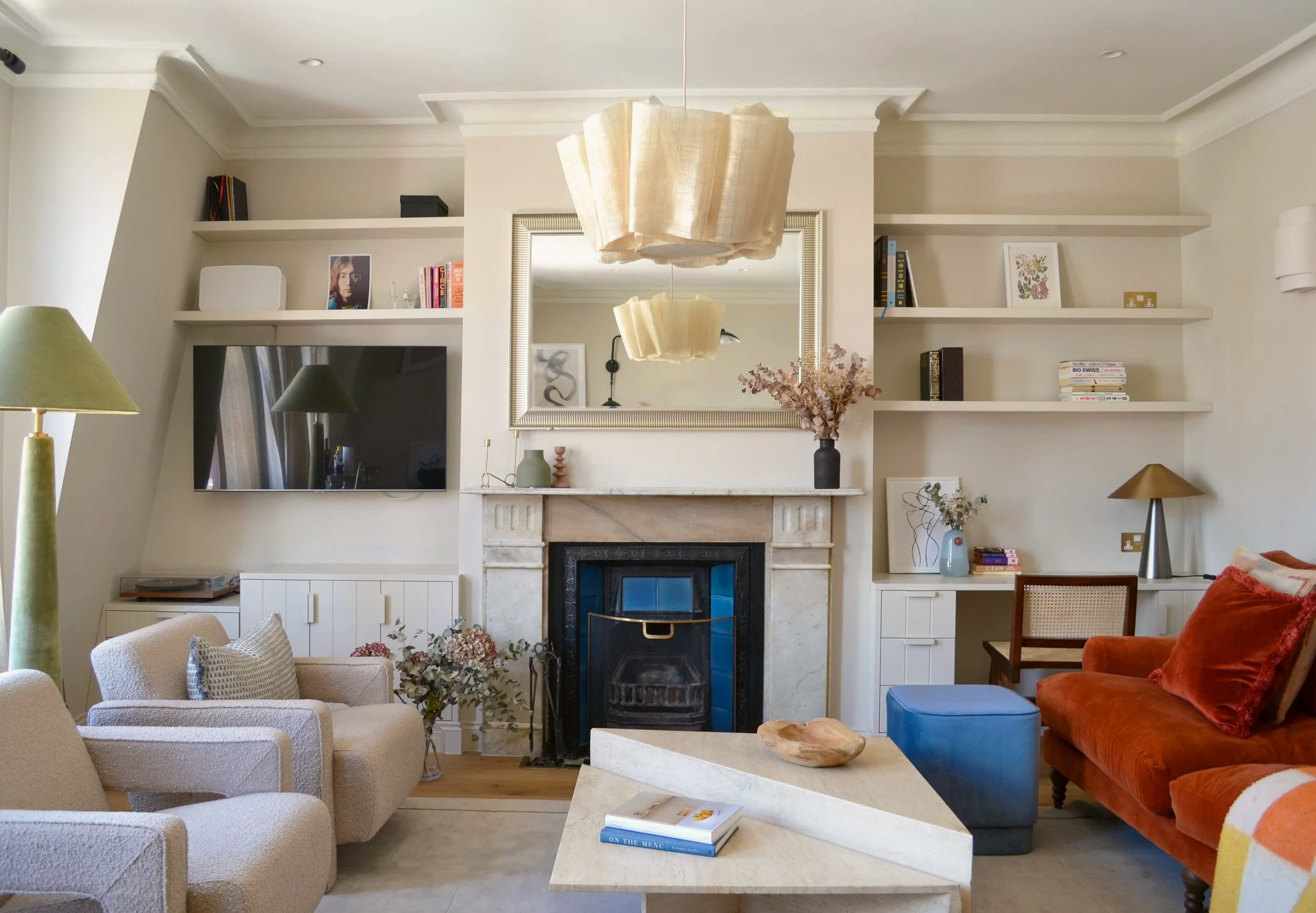 Living room with alcove shelving either side of a marble fireplace, a blue-tiled insert, pleated pendant light, burnt orange sofa and cream armchairs