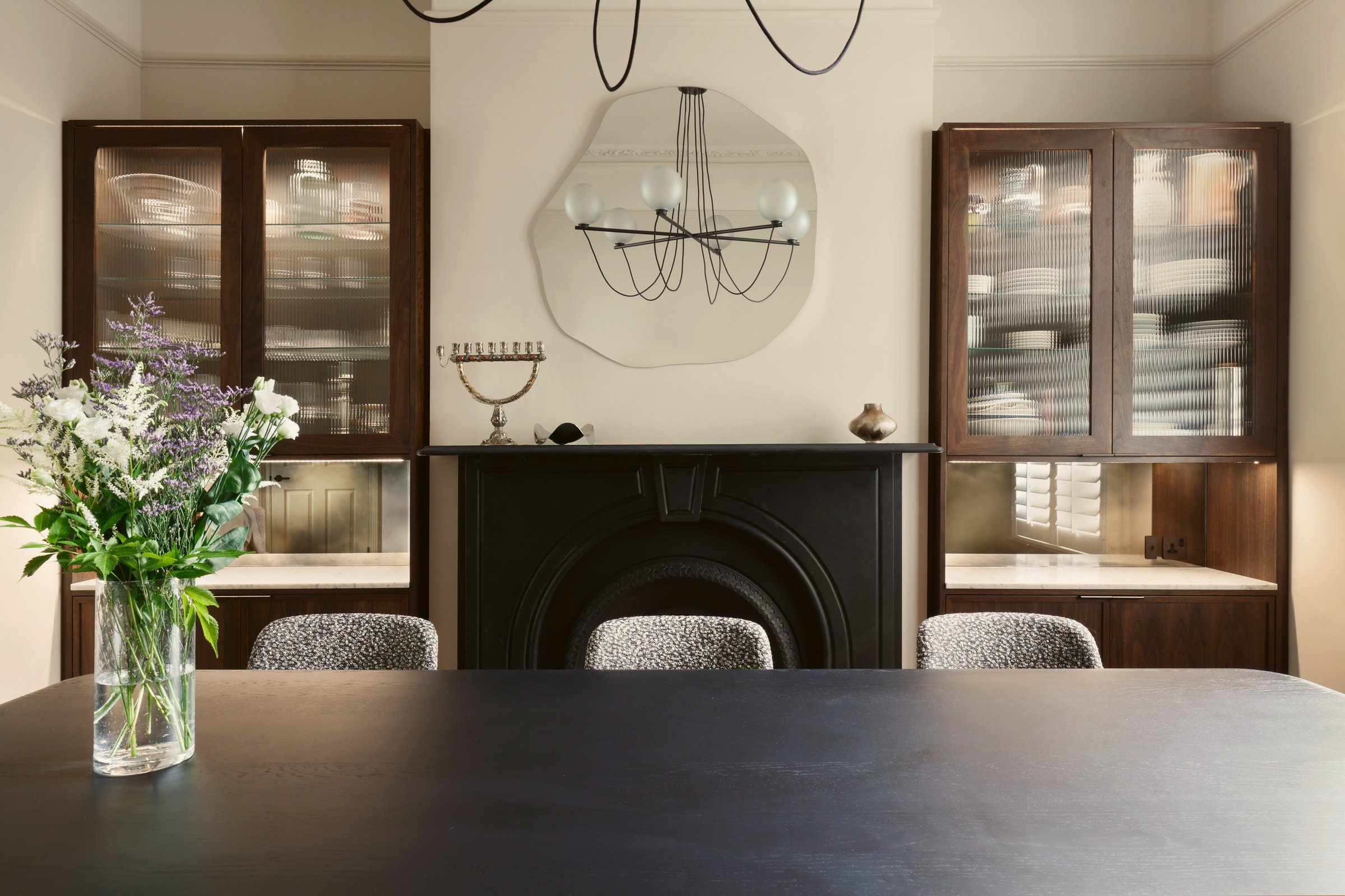 Dining room with a black timber table, a cast-iron fireplace and dark stained dressers holding china behind reeded glass, under an organic-shaped mirror.