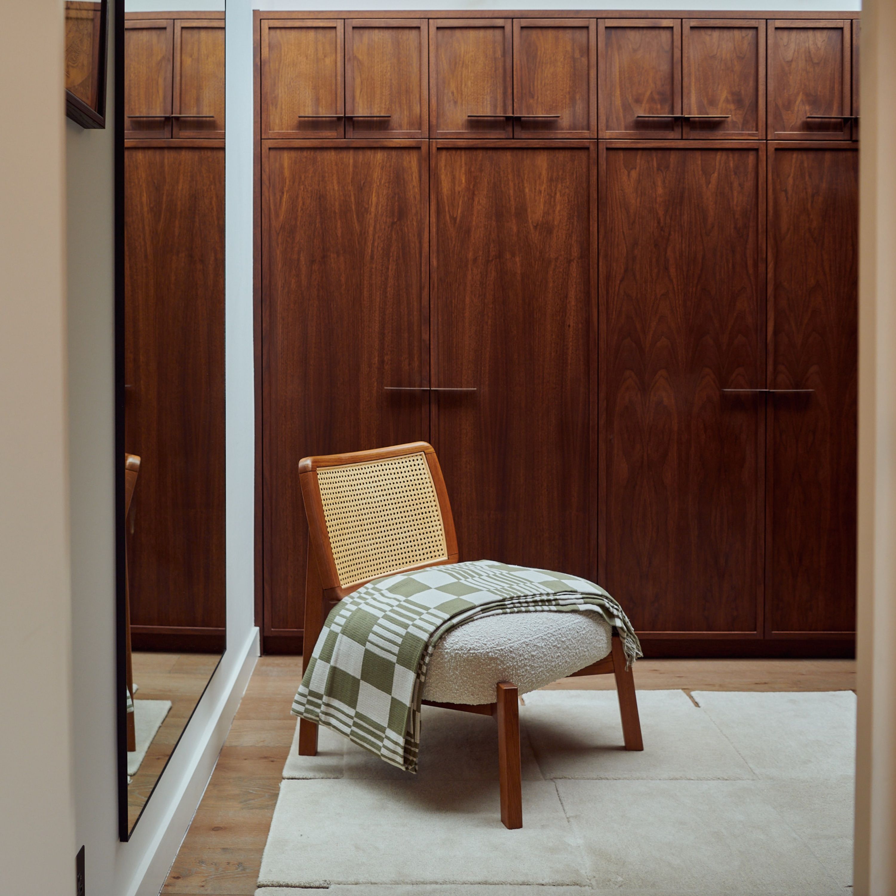 Walnut fitted wardrobes filling a dressing room wall, with a caned lounge chair and green checked throw centred in front and a tall mirror left.