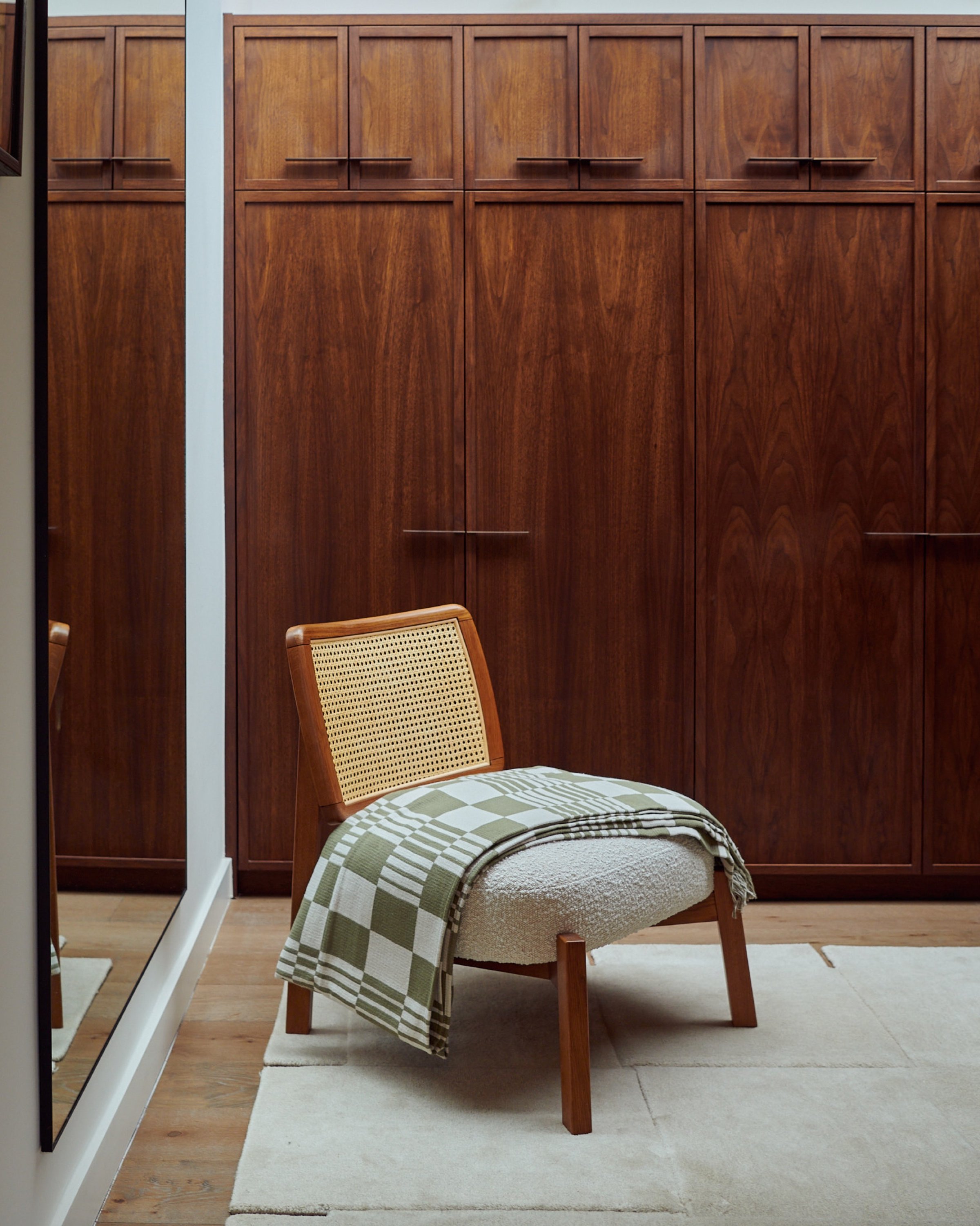 Walnut fitted wardrobes filling a dressing room wall, with a caned lounge chair and green checked throw centred in front and a tall mirror left.