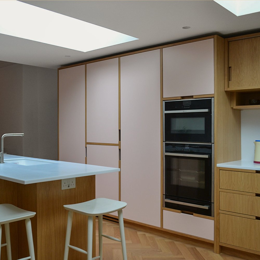 Oak and pale pink kitchen with stacked double ovens, a breakfast bar with stools and a long skylight overhead