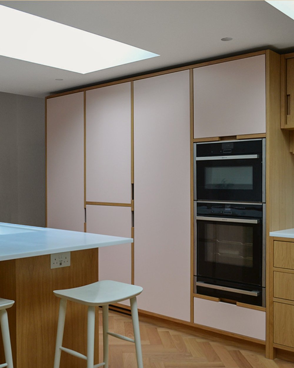 Oak and pale pink kitchen with stacked double ovens, a breakfast bar with stools and a long skylight overhead