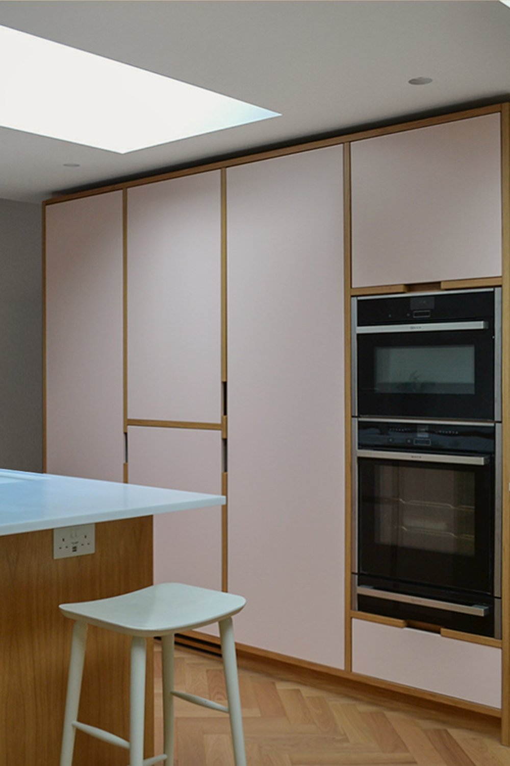 Oak and pale pink kitchen with stacked double ovens, a breakfast bar with stools and a long skylight overhead