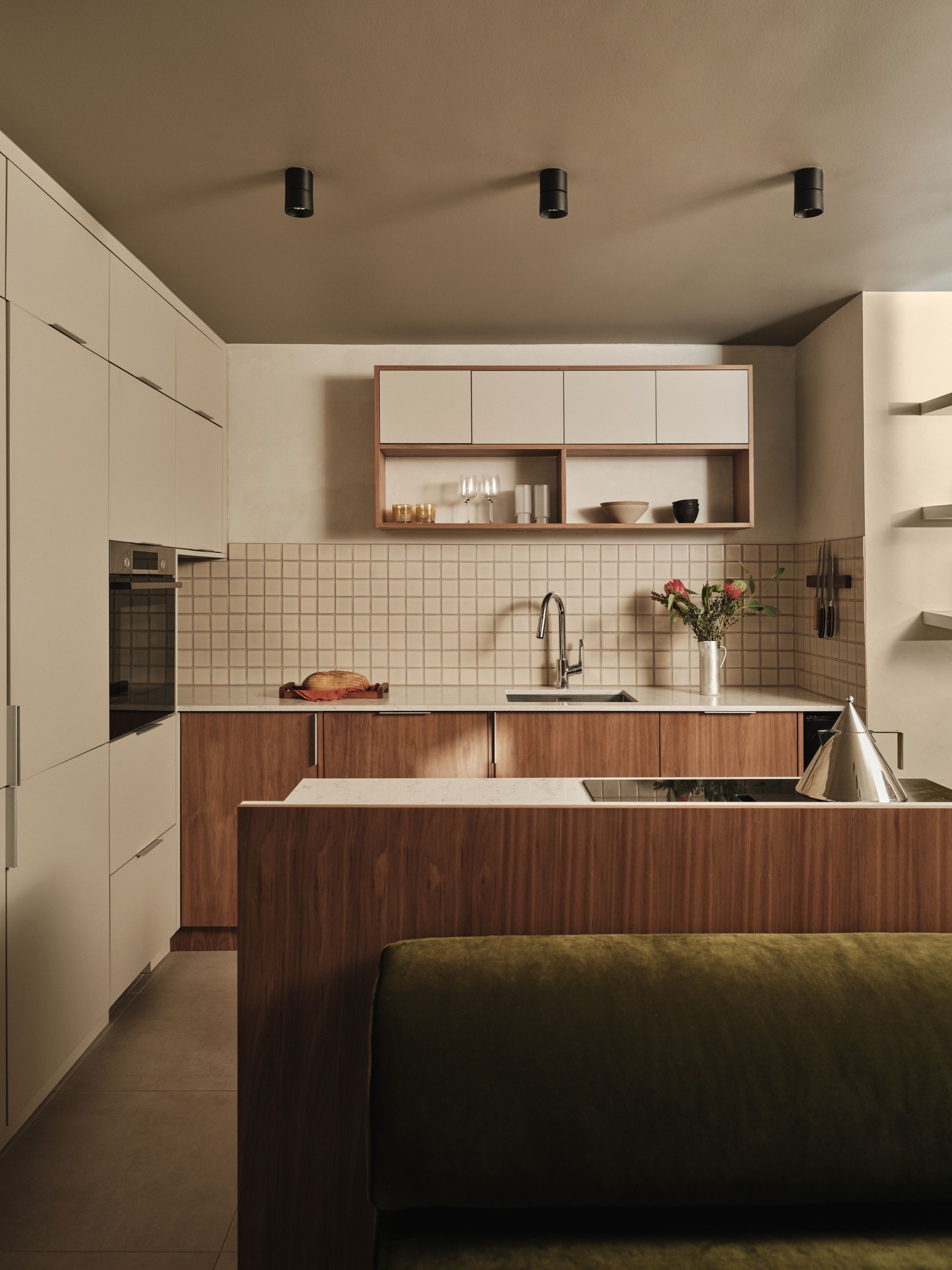 Kitchen with cream handleless tall units, walnut base cabinets under a pale stone worktop, square cream tiling and the olive banquette in front.