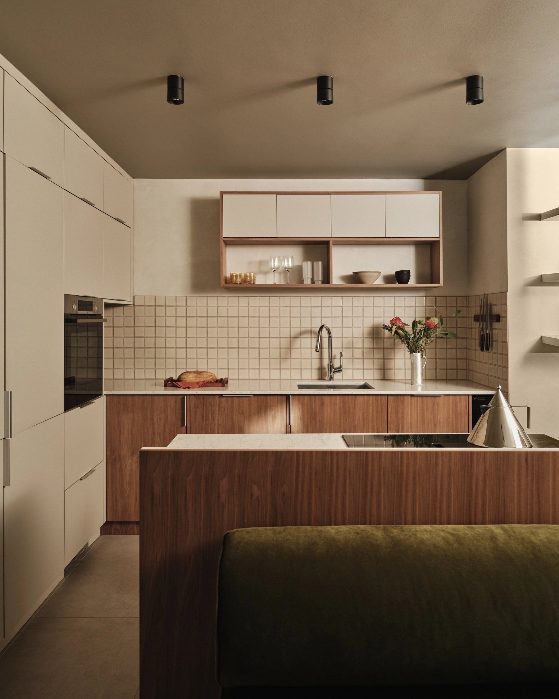 Kitchen with cream handleless tall units, walnut base cabinets under a pale stone worktop, square cream tiling and the olive banquette in front.