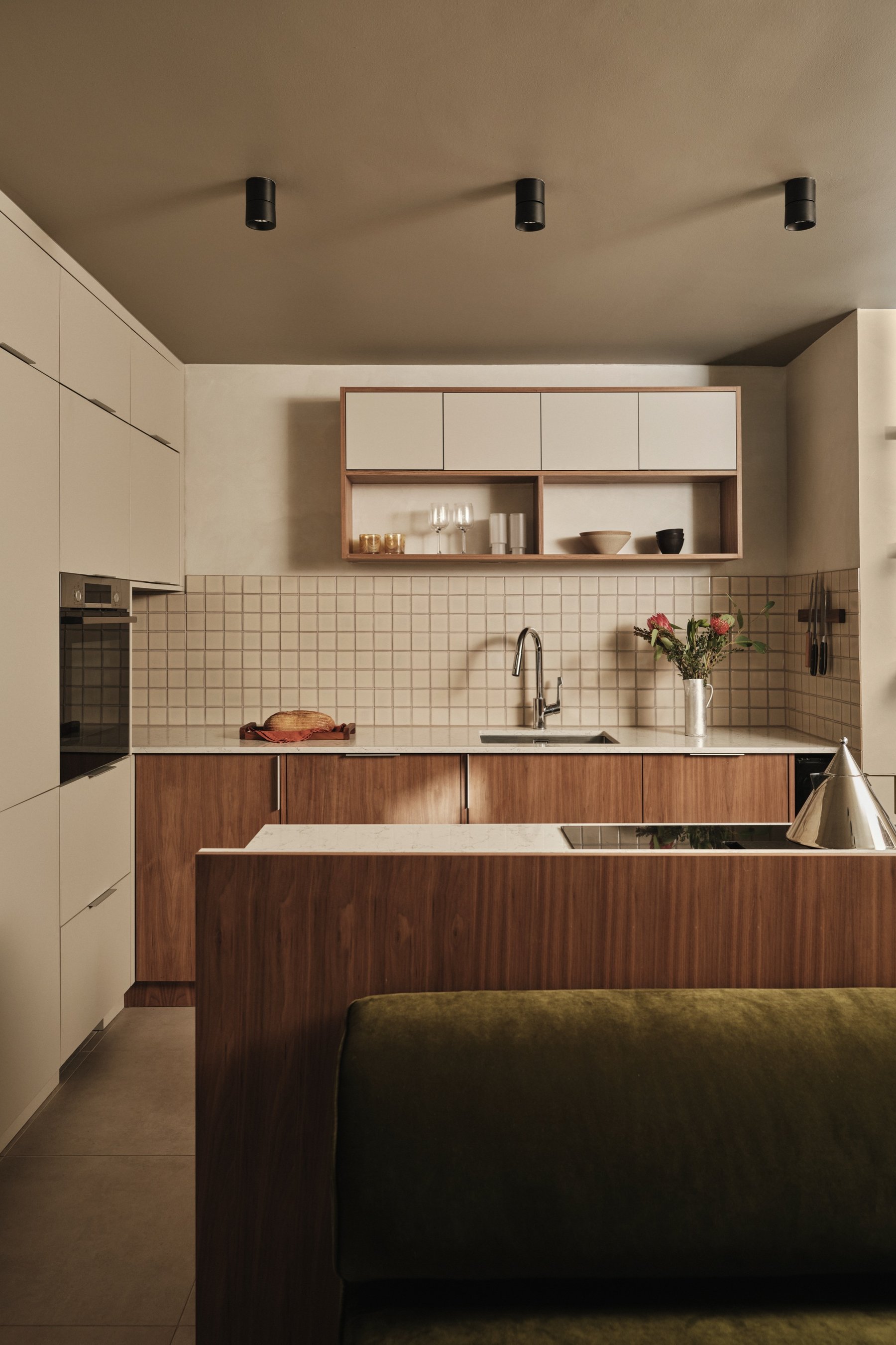 Kitchen with cream handleless tall units, walnut base cabinets under a pale stone worktop, square cream tiling and the olive banquette in front.