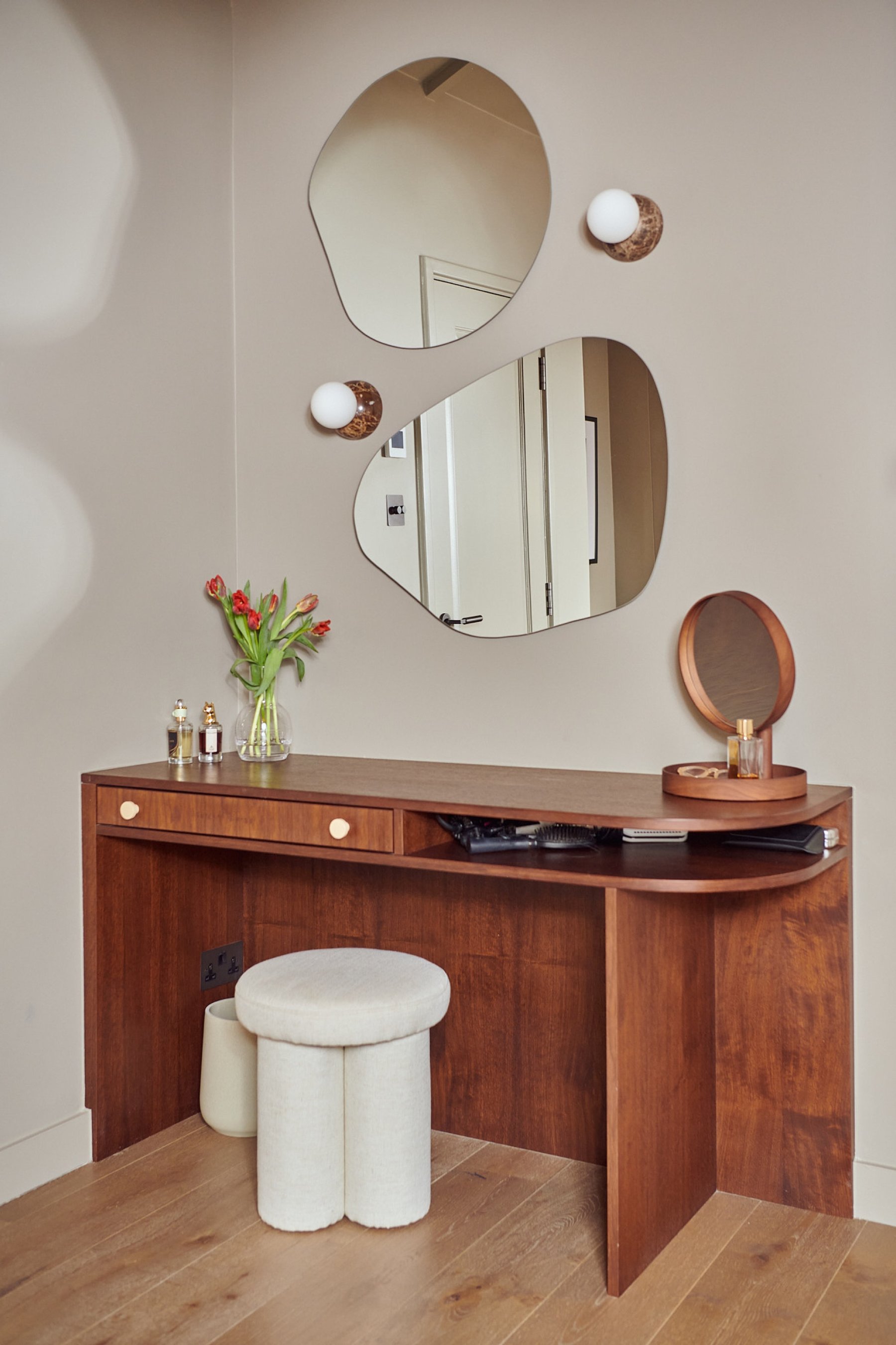 Walnut dressing table with a curved end, two organic-shaped mirrors and small globe wall lights above, and a boucle stool tucked beneath.