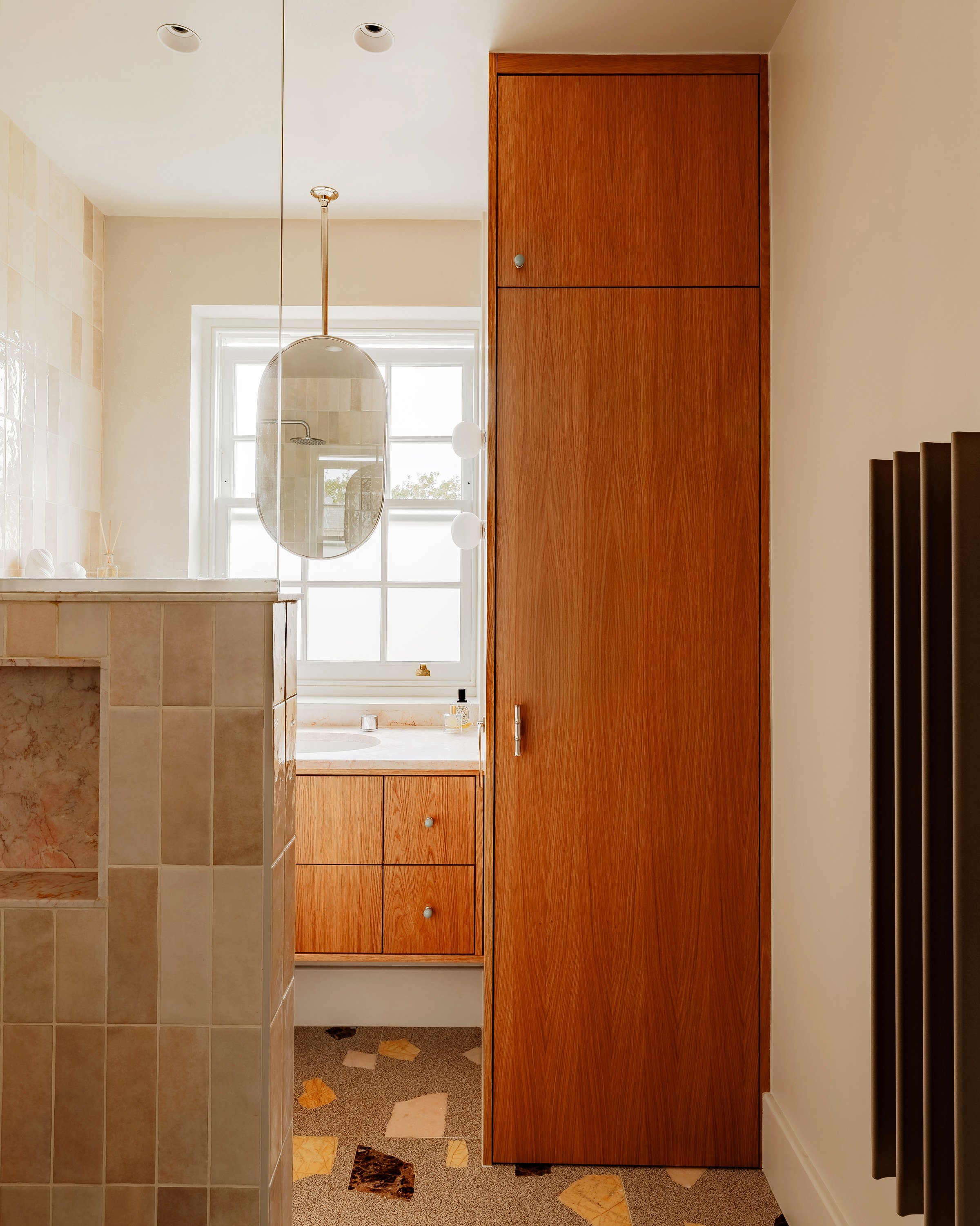 A tall wooden cabinet beside the vanity and the tiled shower wall, with a pendant mirror at the sash window.