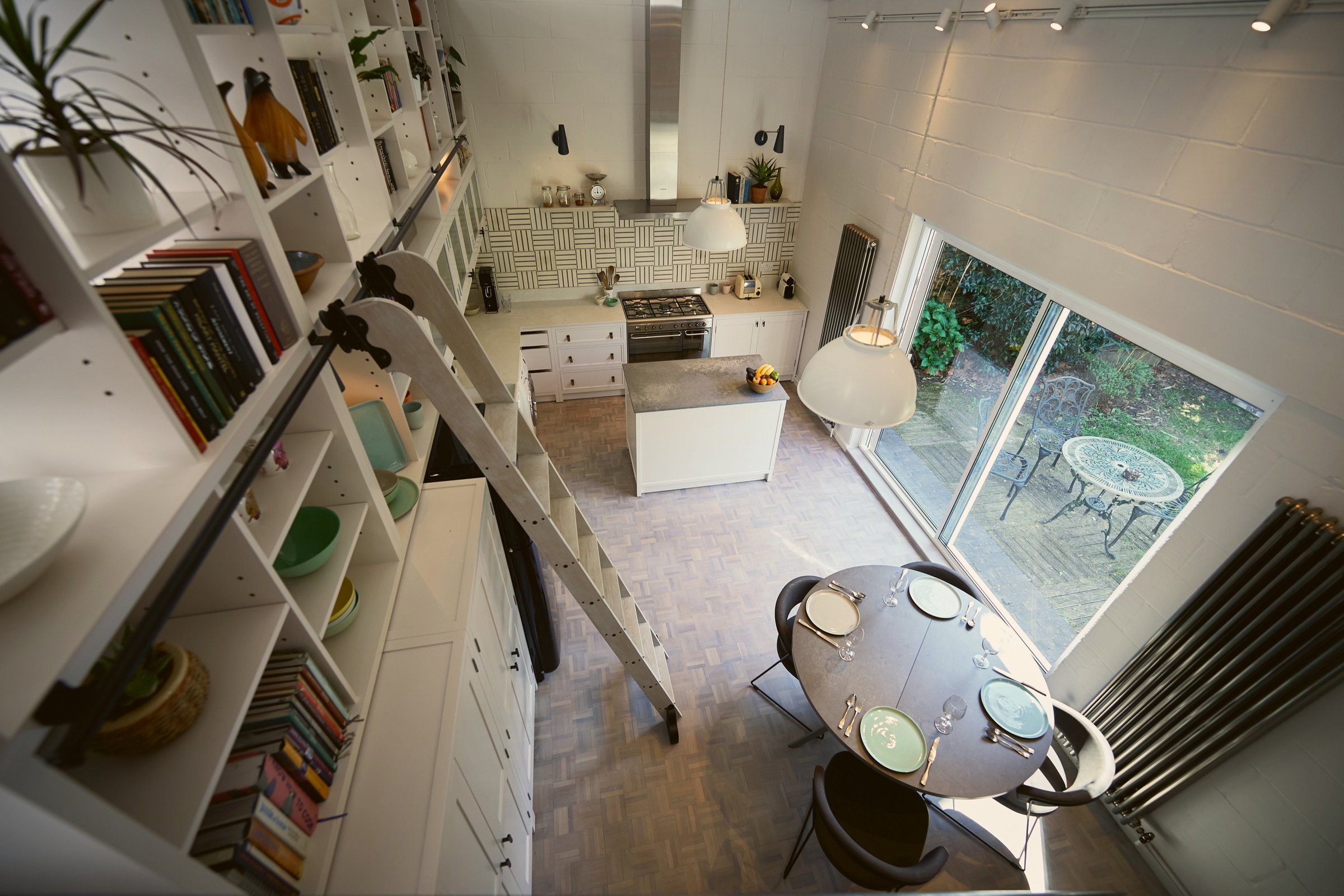 Looking down into the kitchen from the mezzanine: white joinery and library ladder left, island and range below, round table laid by the garden doors.