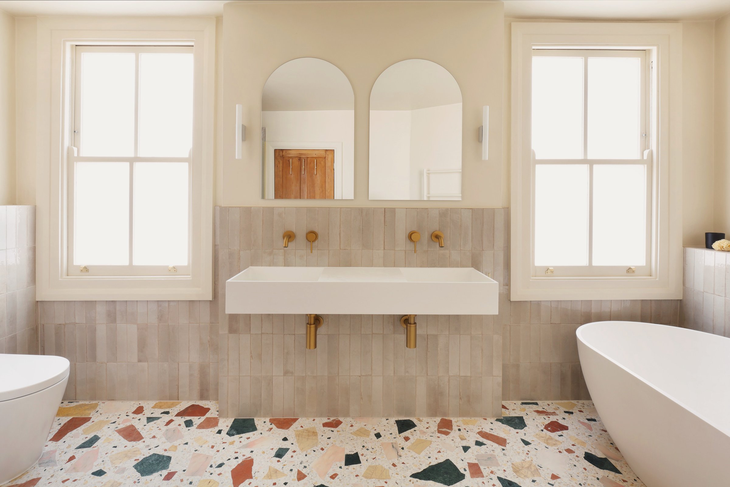 Wide view of the bathroom: twin-mirrored trough basin between two sash windows, freestanding bath at the right, all over large-chip terrazzo.