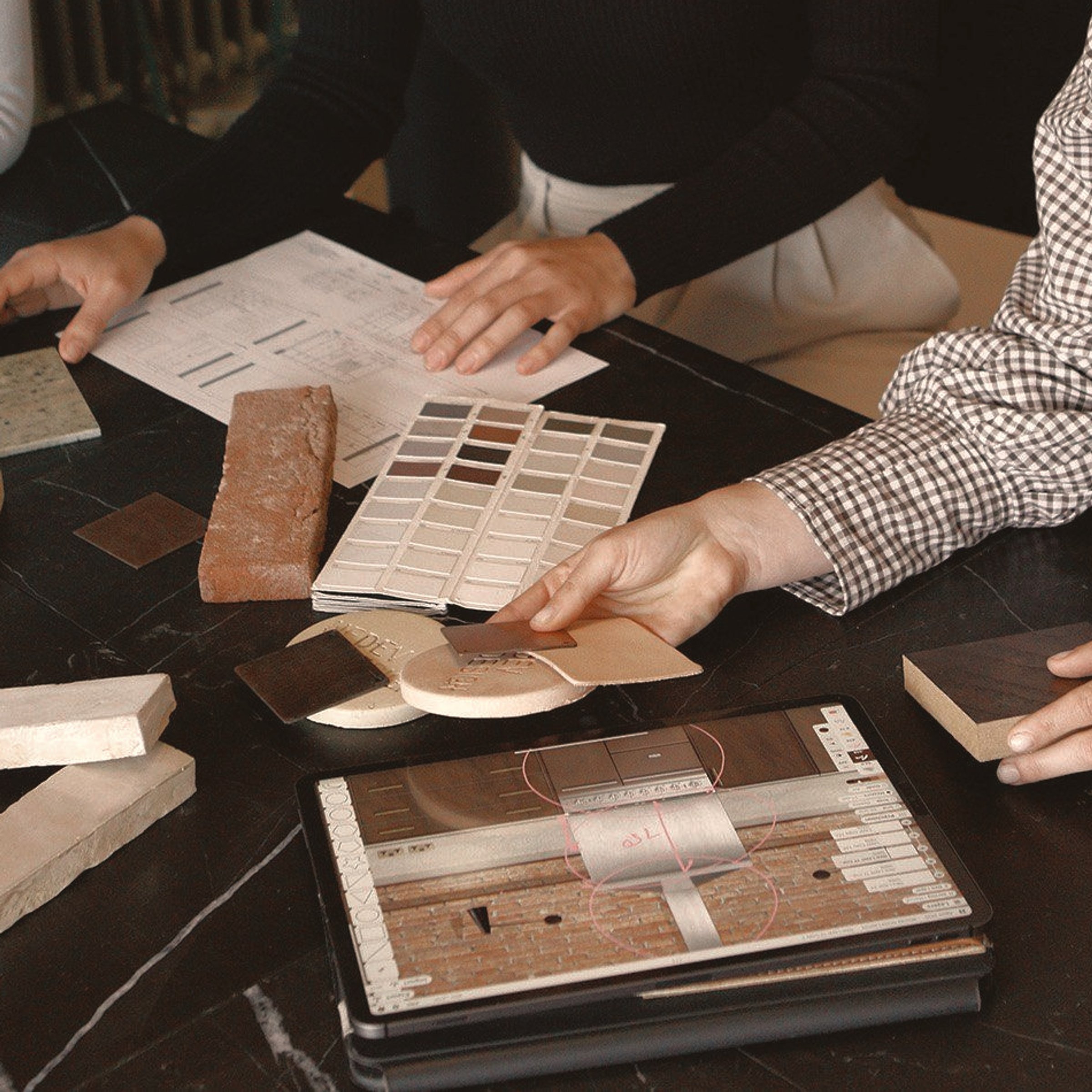 Three designers at a dark marble table, working through stone, brick, tile and fabric samples beside a printed palette.