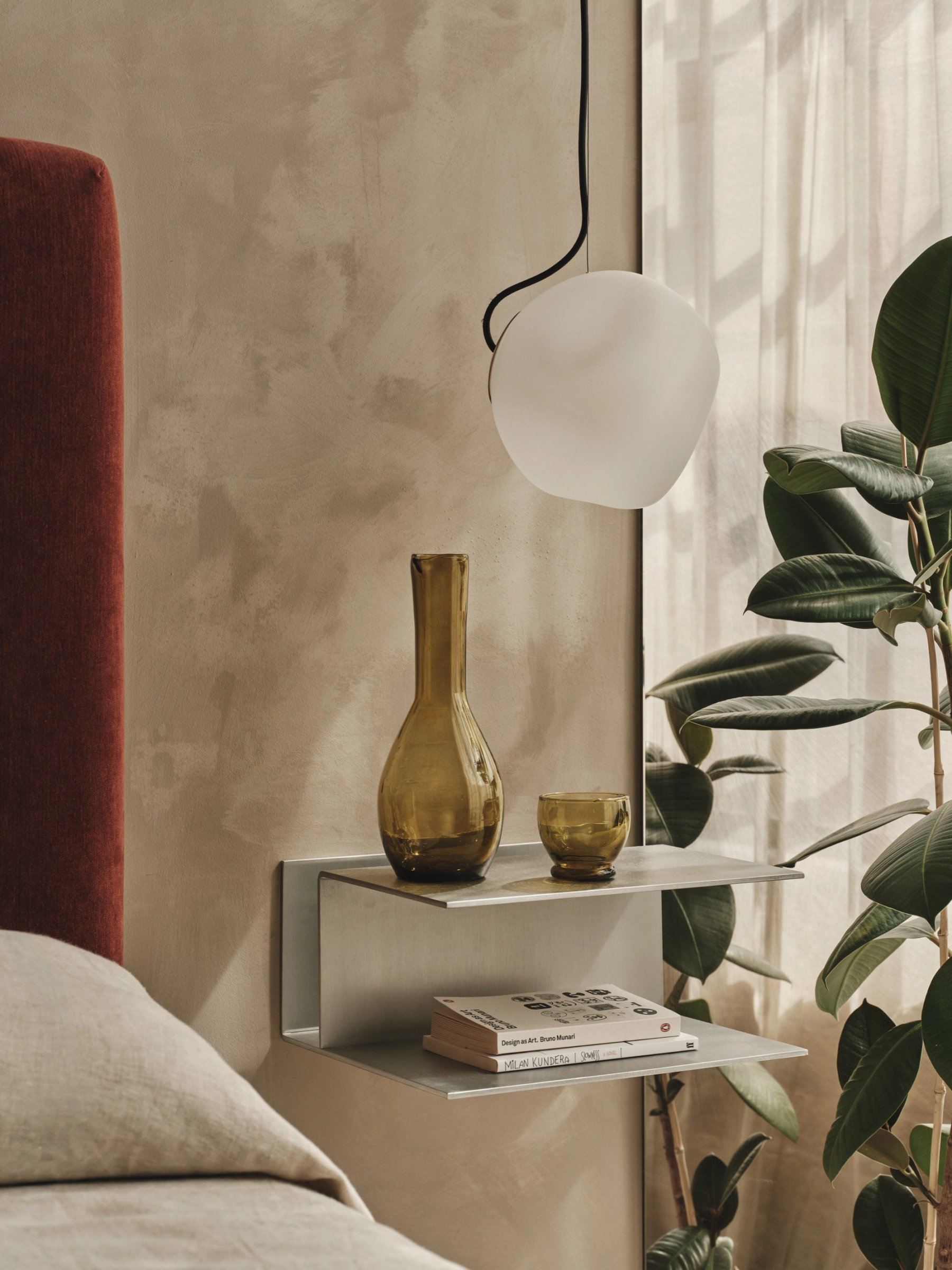 Bedside detail: a folded metal shelf carrying an amber glass carafe over a stack of books, with a white globe pendant hanging beside it.