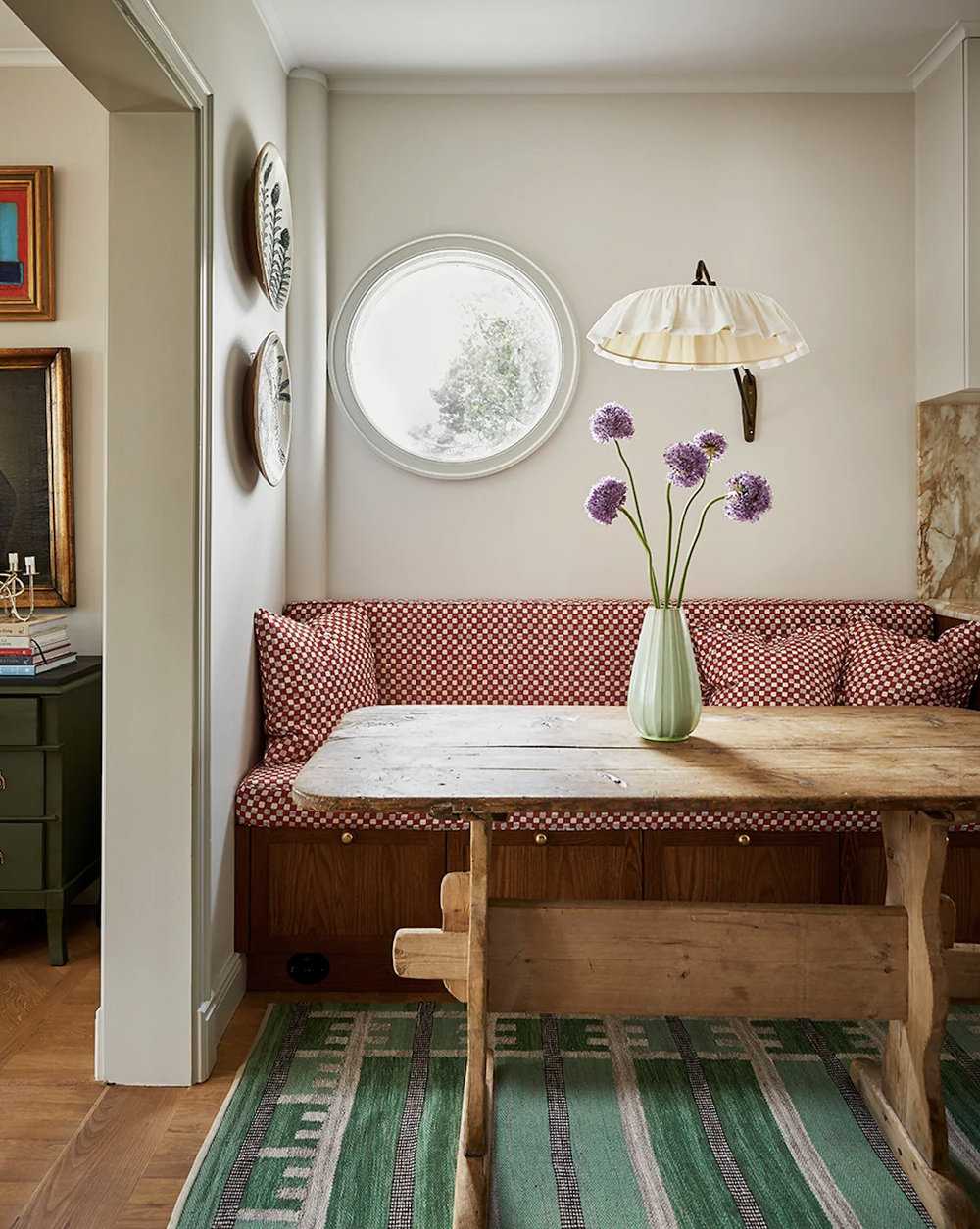 Dining nook with a built-in banquette upholstered in a red and white check, a rustic wooden trestle table and a green striped rug, beneath a round window