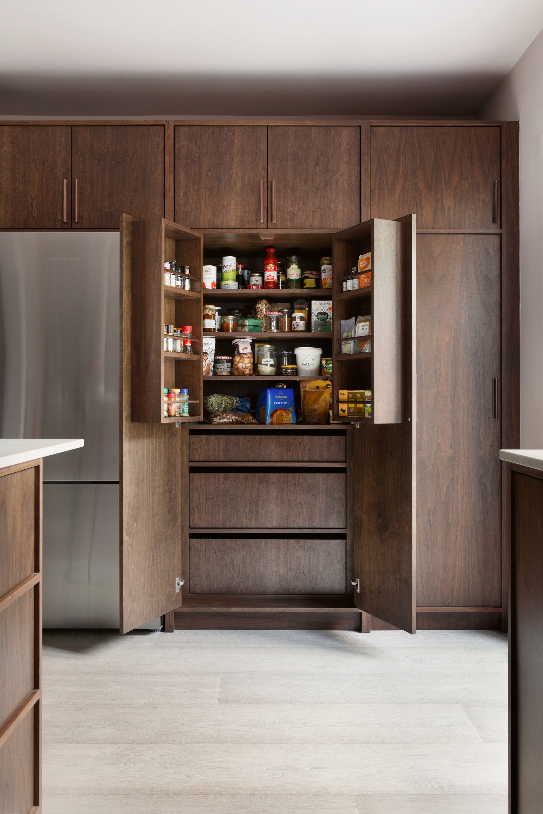 The open larder in its run of dark timber cupboards, shelves and door racks full, with the fridge left and the island worktop in front.