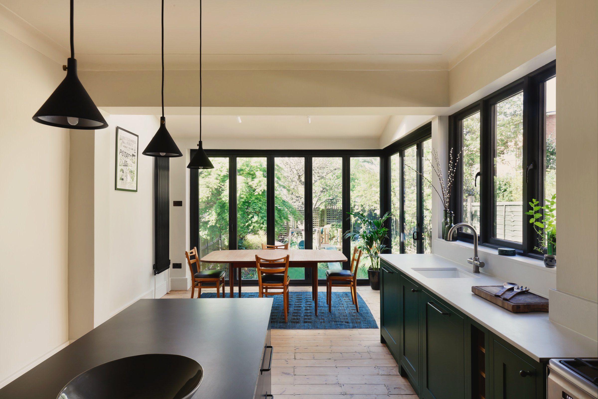 Kitchen looking down the room to a rear extension glazed in black steel, three black cone pendants over a dark stone island and green cabinetry at the right.