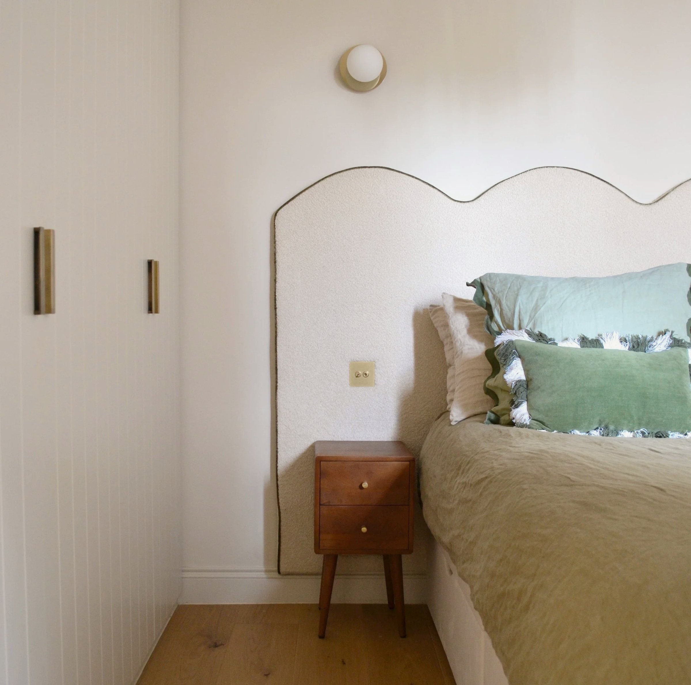 Bedroom with a shaped upholstered headboard in cream linen, sage green and blush pillows, and a small two-drawer walnut bedside table on an oak floor