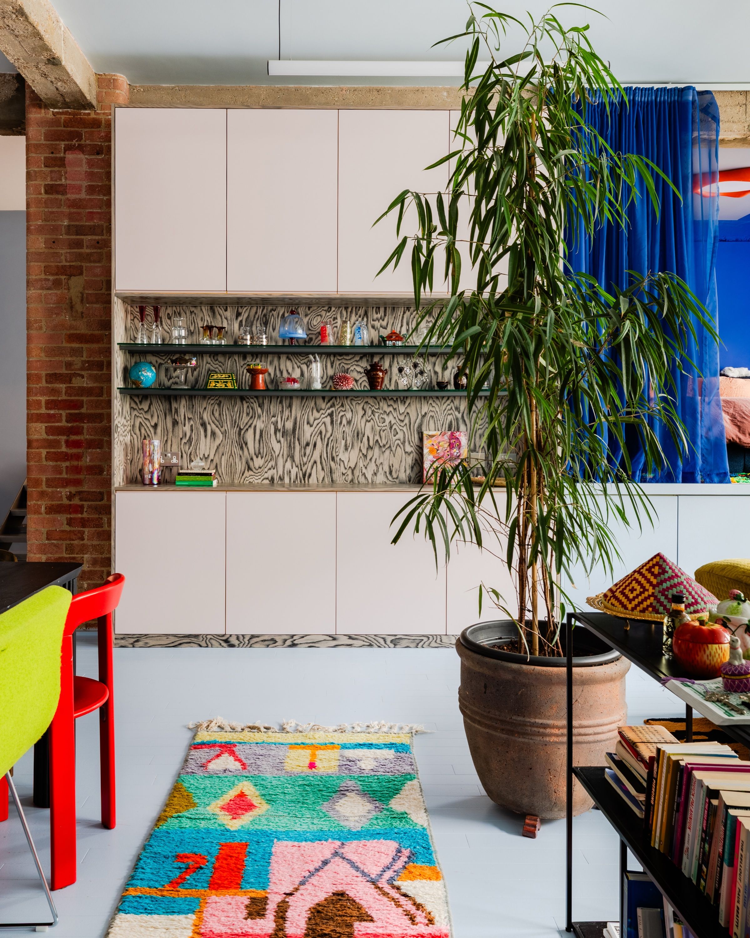 Pink fitted cabinetry with a figured ply shelf recess, over pale blue painted boards, with a Berber rug and a bamboo in a terracotta pot in front.