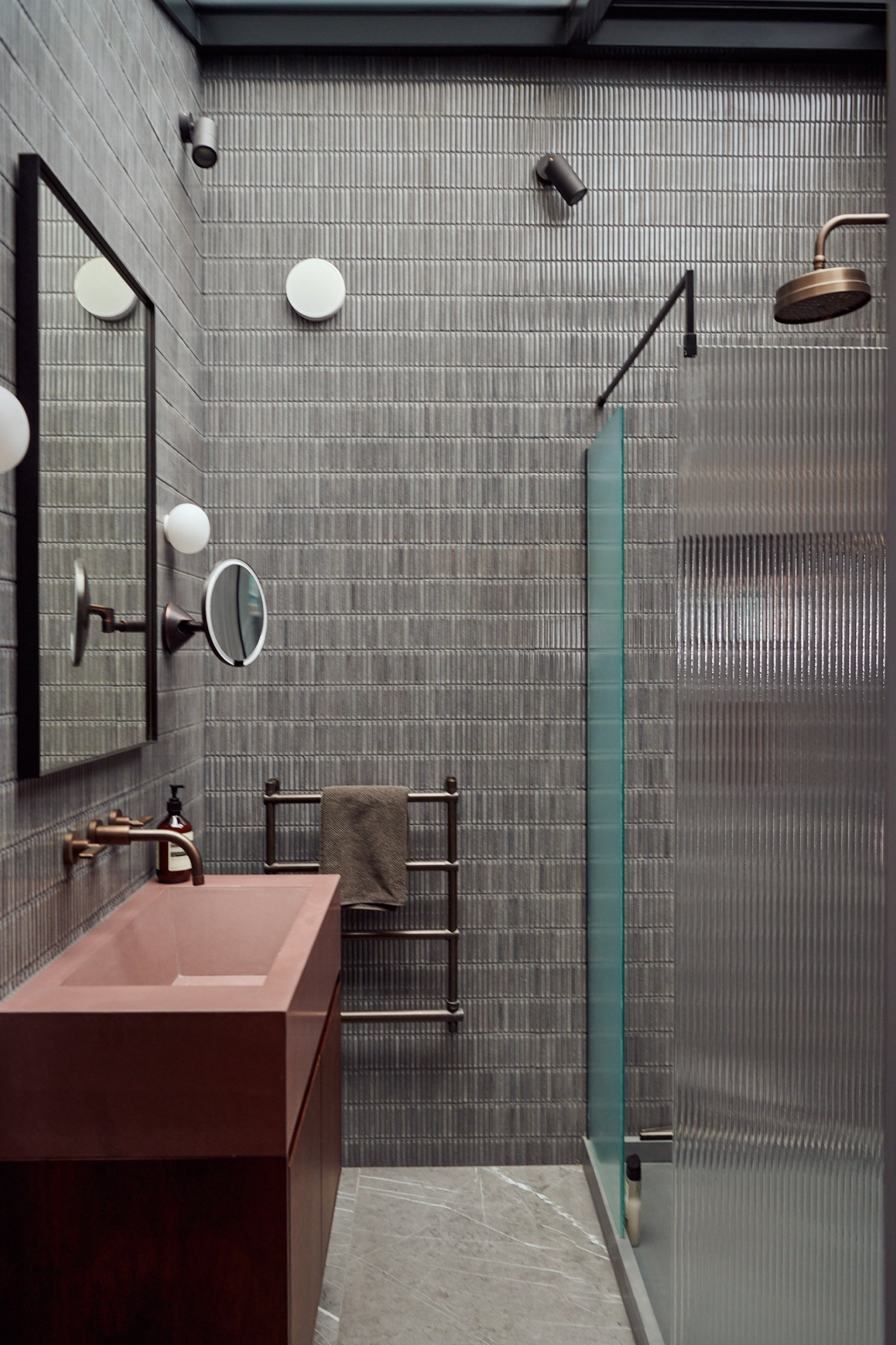 Shower room lined in narrow grey tile, with a rust trough basin on a timber vanity, aged brass fittings and a reeded glass screen.