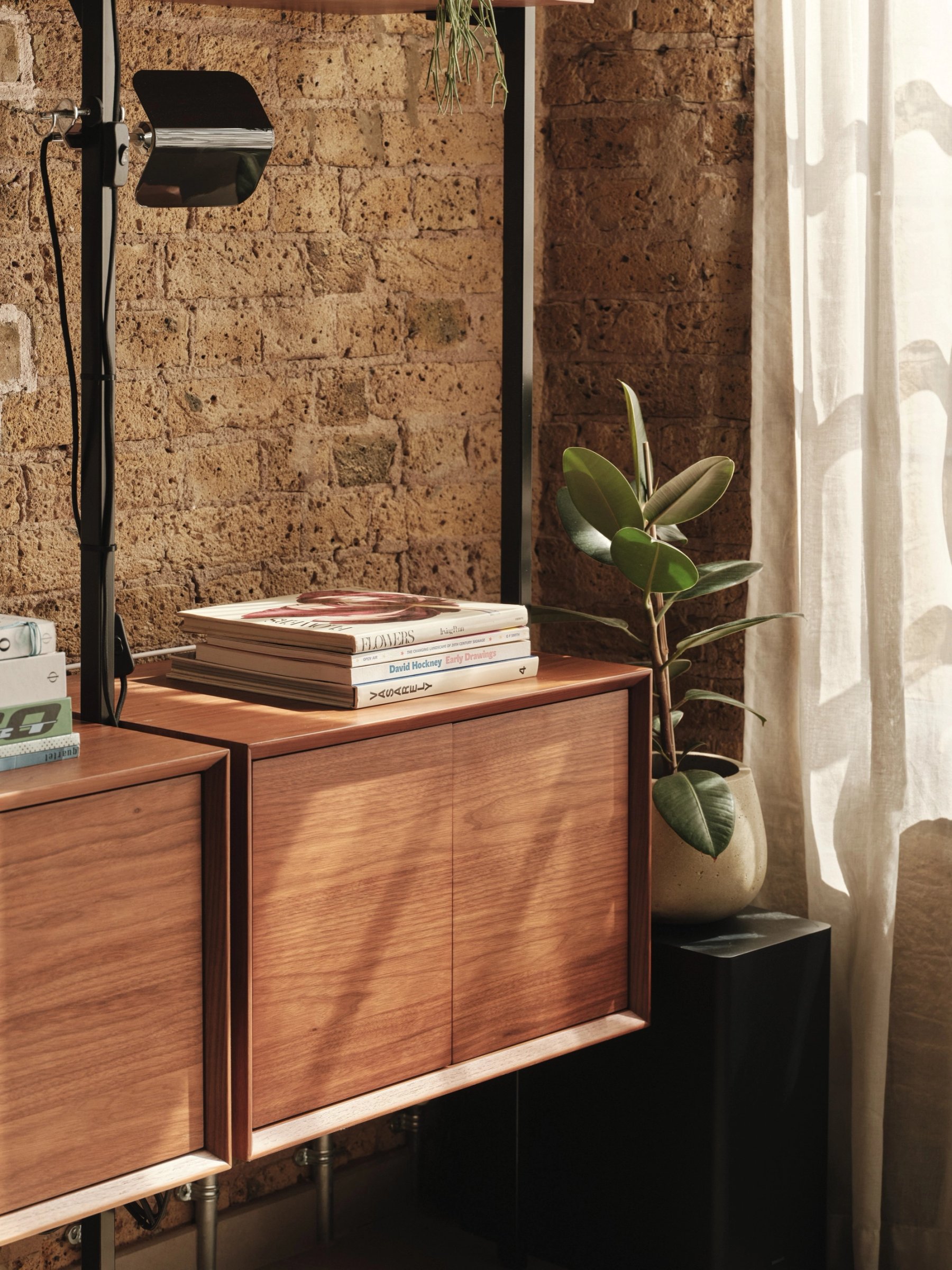 Detail of the wall-hung teak cabinets against exposed brick, books stacked on top in low light, with an adjustable black wall lamp above.