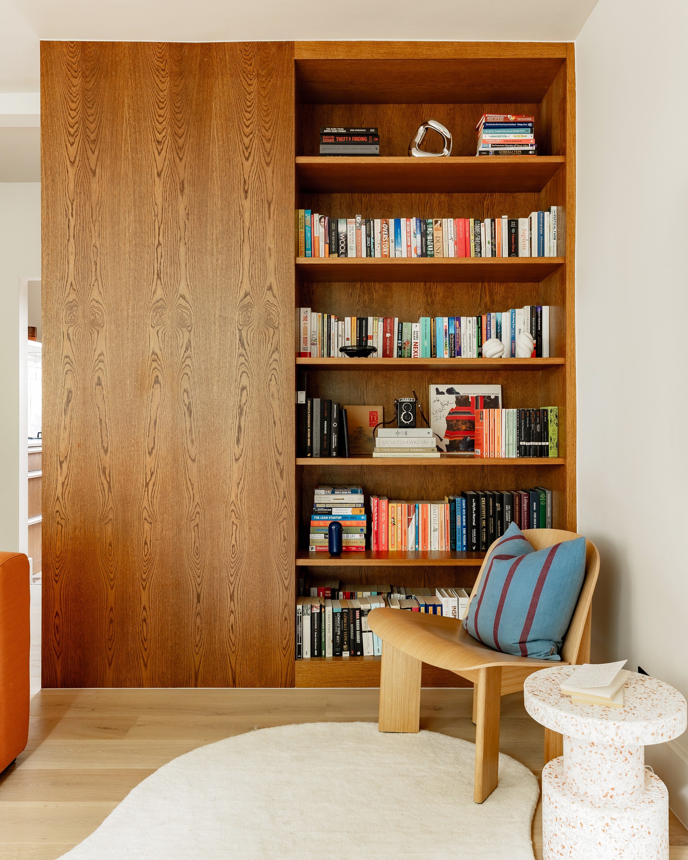 A wall of wood joinery in the living room: a full-height panelled cabinet beside open shelves of books, with an armchair and a white side table in front.