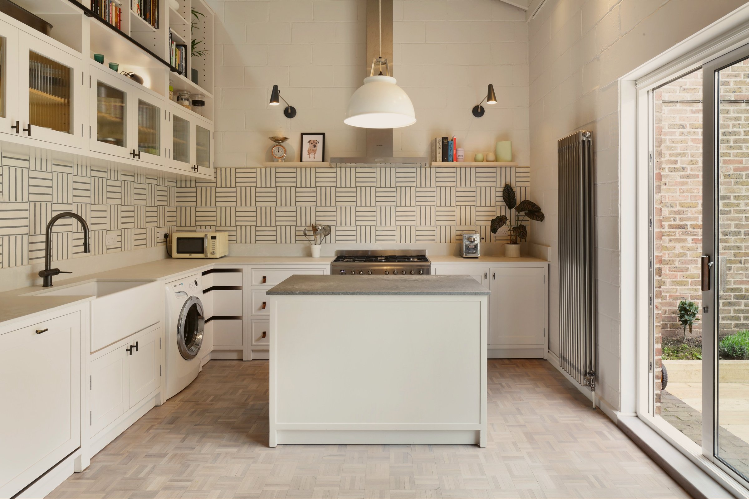 White kitchen in a double-height room, with a monochrome basketweave splashback, glazed wall cupboards, a small island and full-height garden doors.