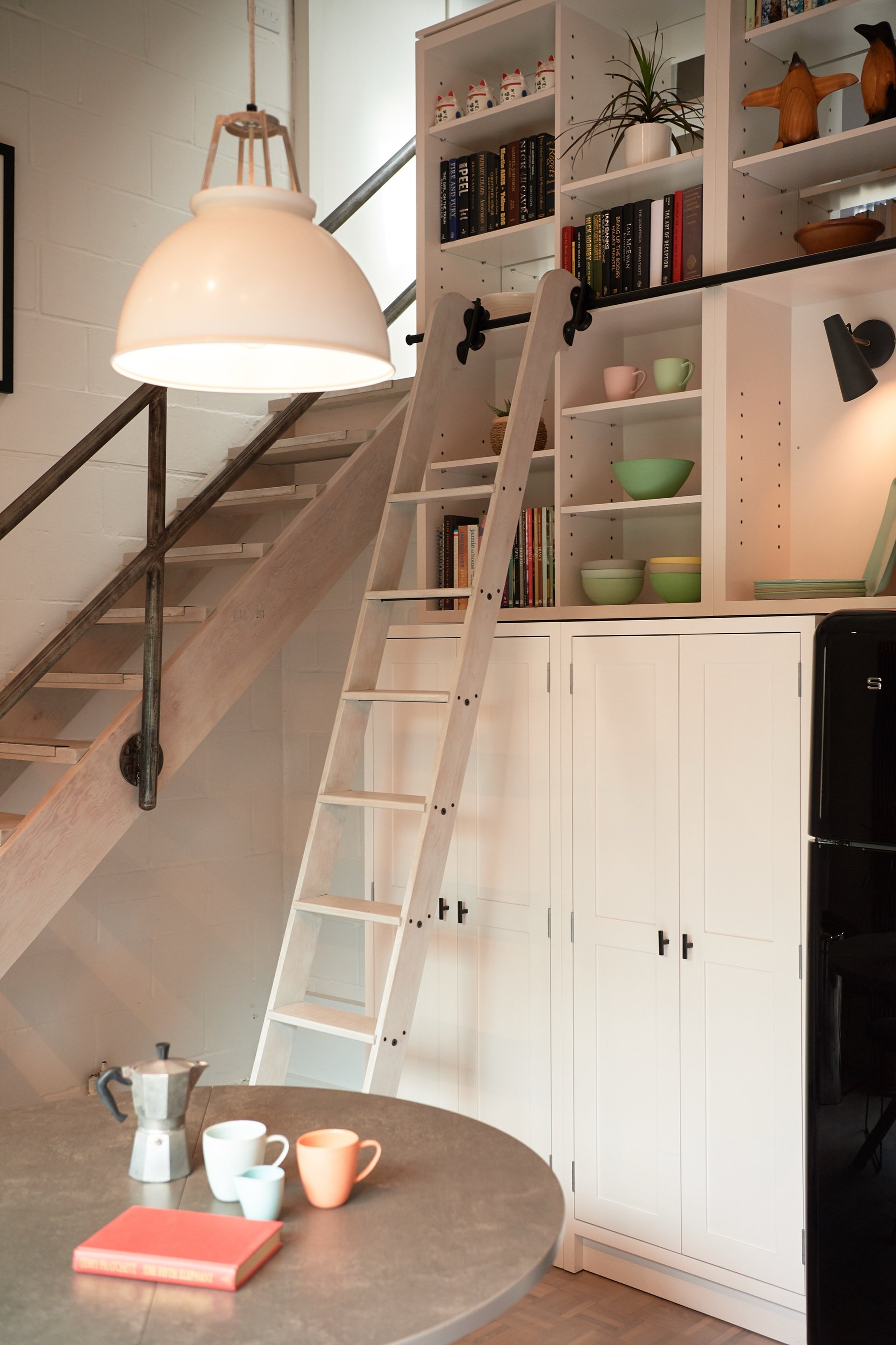 Kitchen under the open stair, with a library ladder on its rail against white shelving, a black fridge right and a white pendant over a round table.