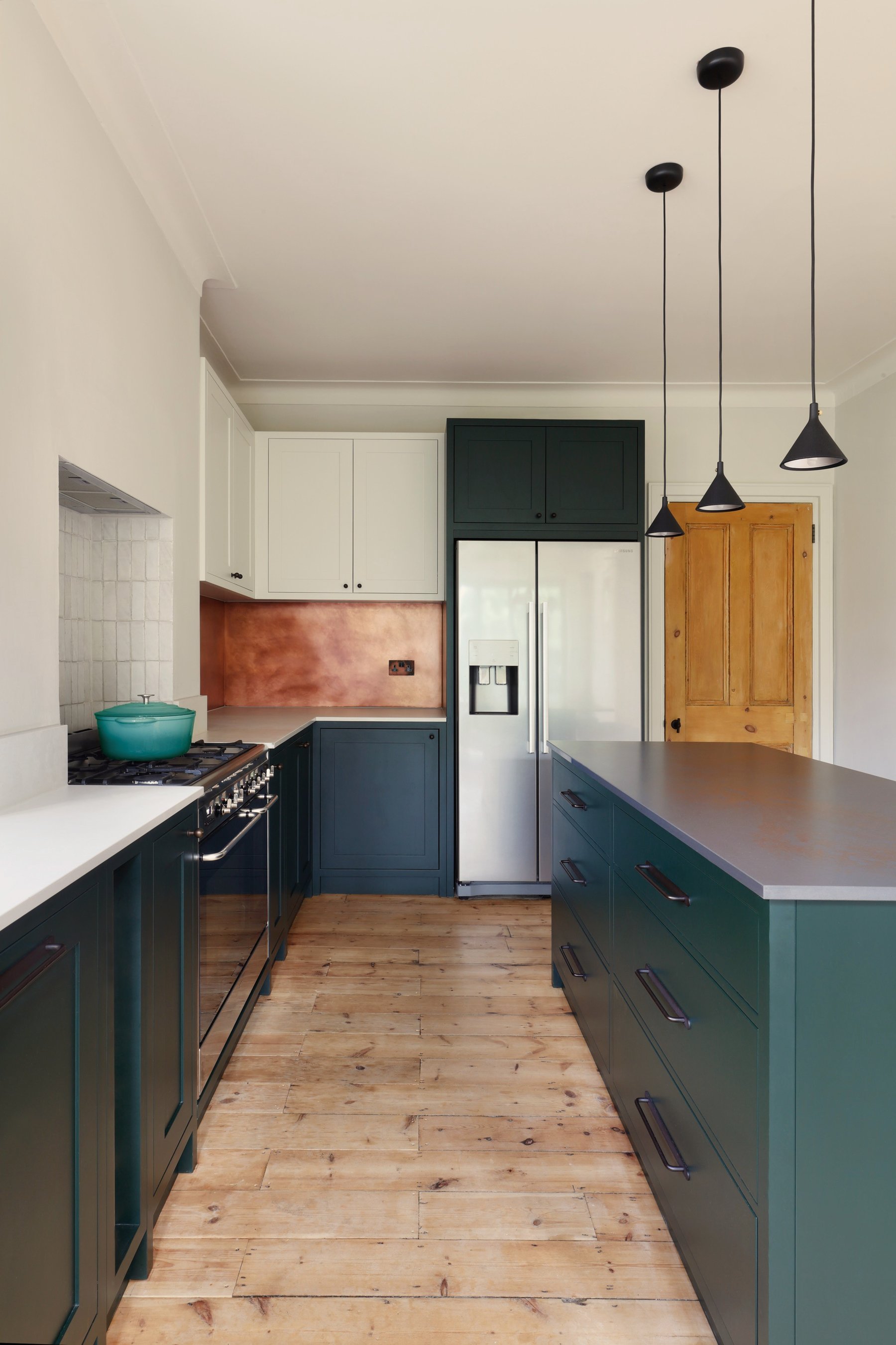 Galley view with dark green cabinets both sides, a stone-topped island running towards the camera and the copper splashback at the far end.