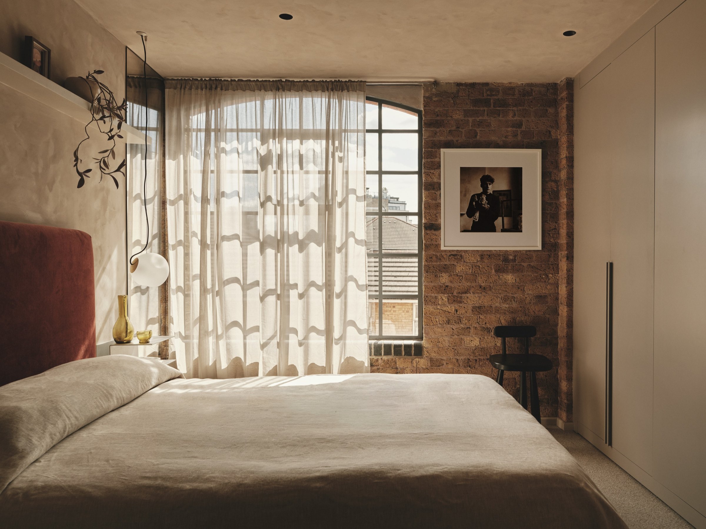 Warehouse bedroom facing a tall arched steel window hung with sheer curtains, a rust velvet headboard against polished plaster and exposed brick beyond.