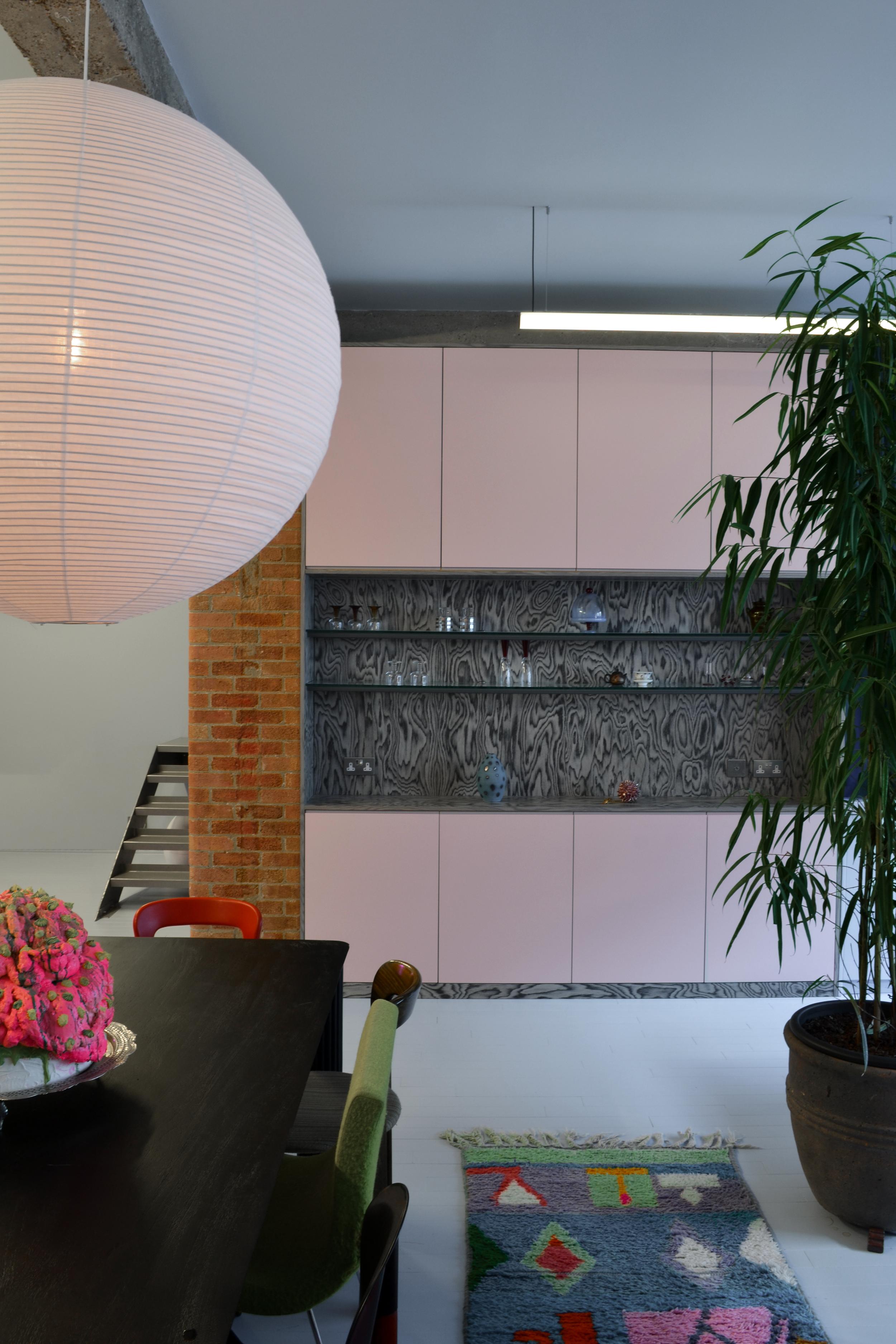 Dining area under a large pink paper globe, with pale pink cabinets, an antiqued mirror shelf of glassware and an exposed brick pier