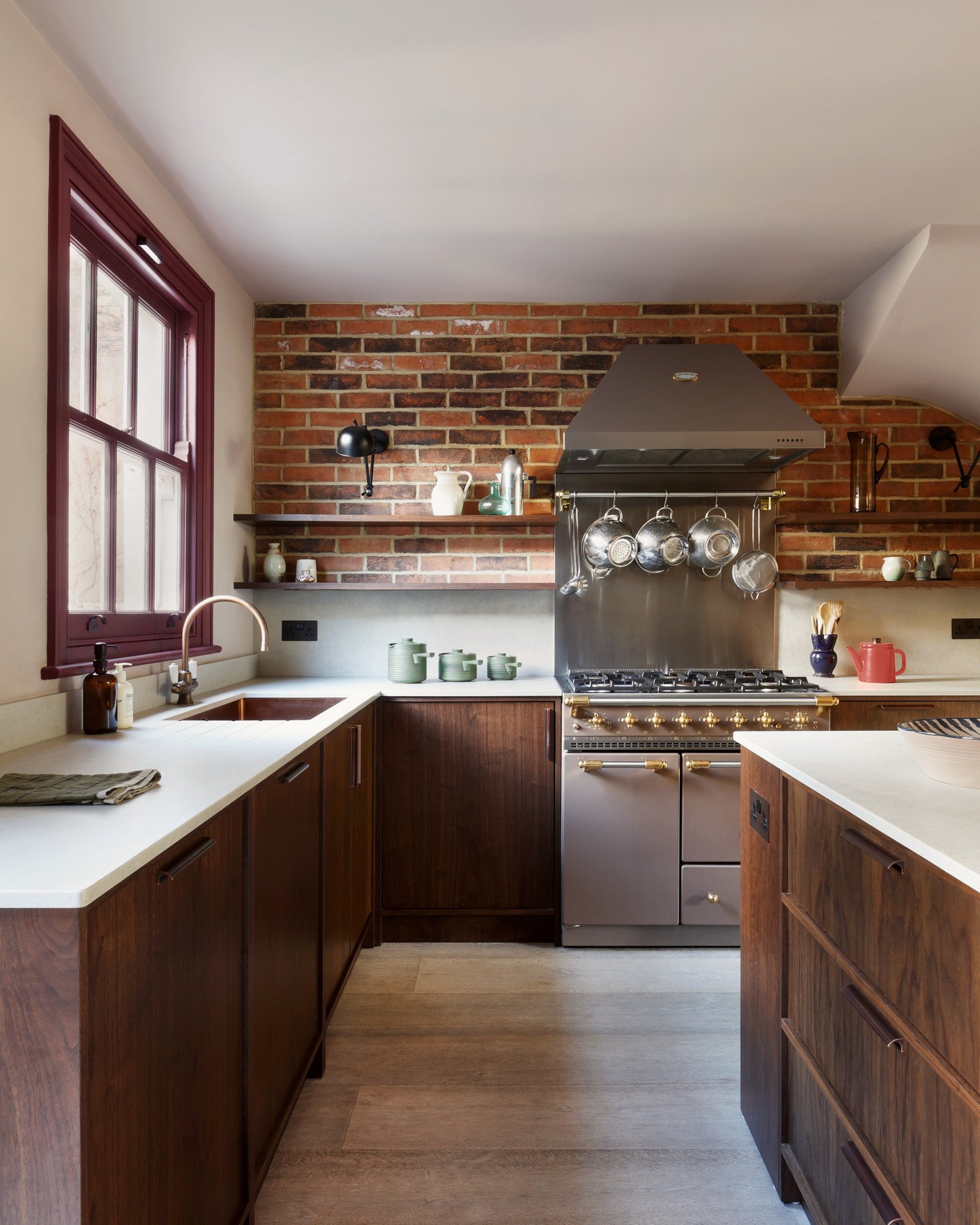 Kitchen with dark stained timber cabinets both sides, a stainless range against an exposed brick chimney breast and copper pans on a rail.