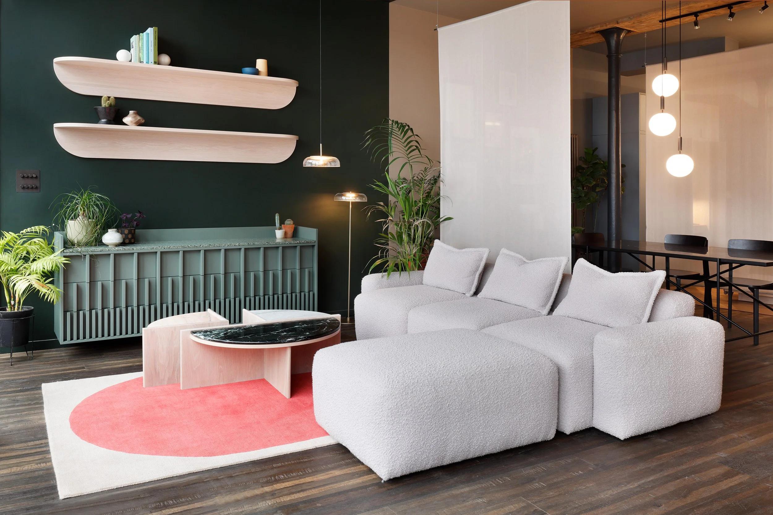 Showroom with a dark green wall, two curved pink floating shelves above a ribbed green sideboard, and a white boucle modular sofa on a round pink rug