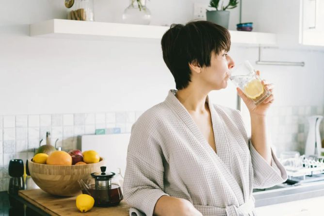 Žena v županu pije ráno vodu s plátkem citronu v útulné kuchyni
