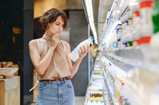 Žena zamyšleně vybírá lahvičku kefíru v supermarketu mezi ostatními mléčnými výrobky Žena zamyšleně vybírá lahvičku kefíru v supermarketu mezi ostatními mléčnými výrobky